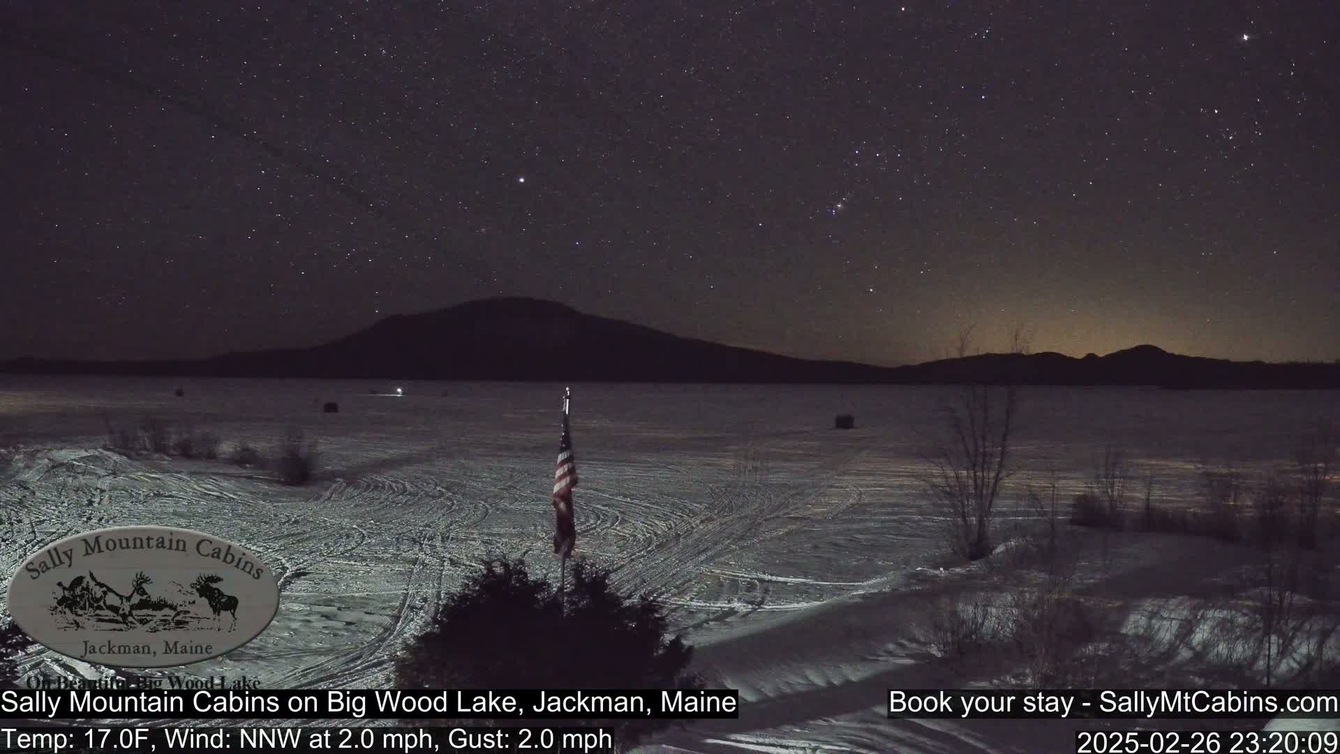A starry night sky over a snow-covered frozen lake with a silhouetted mountain in the background, under a 17°F temperature with a light north-northwest wind.