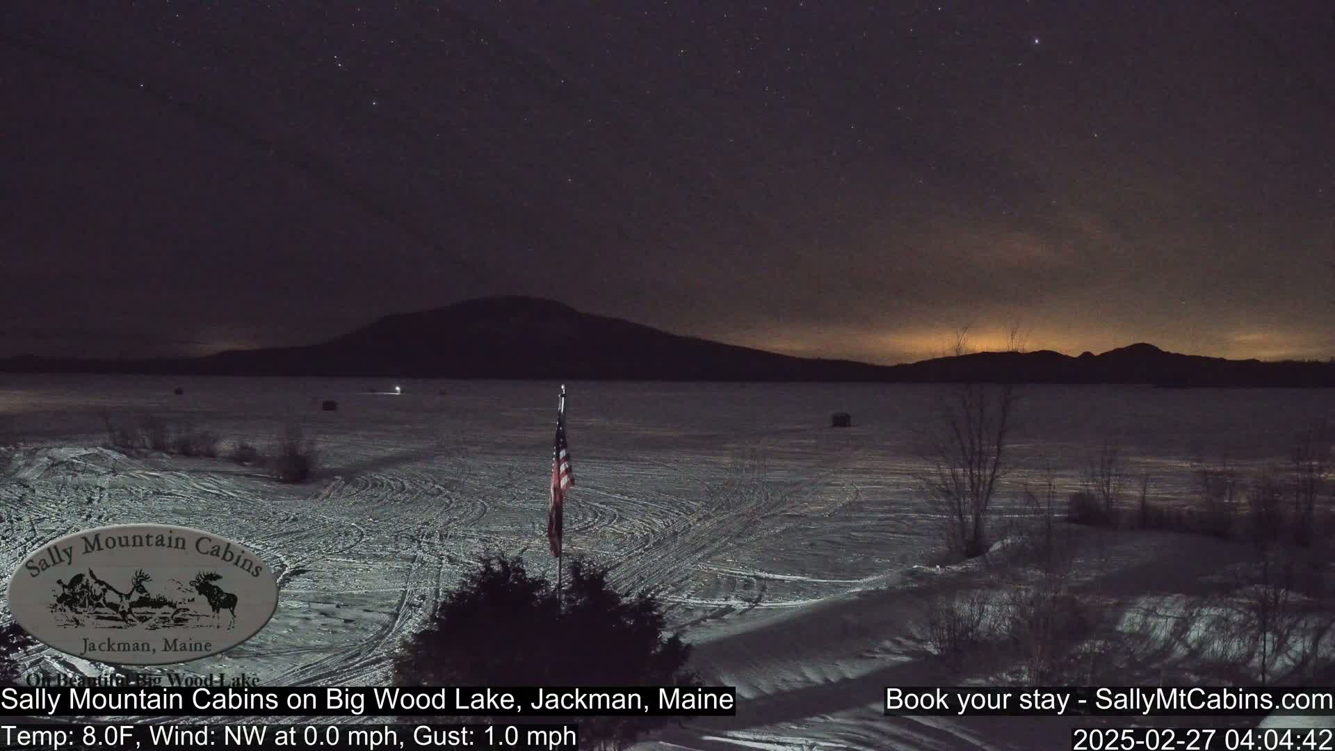 A frozen lake under a starry night sky, with a mountain range in the background and an American flag in the foreground.