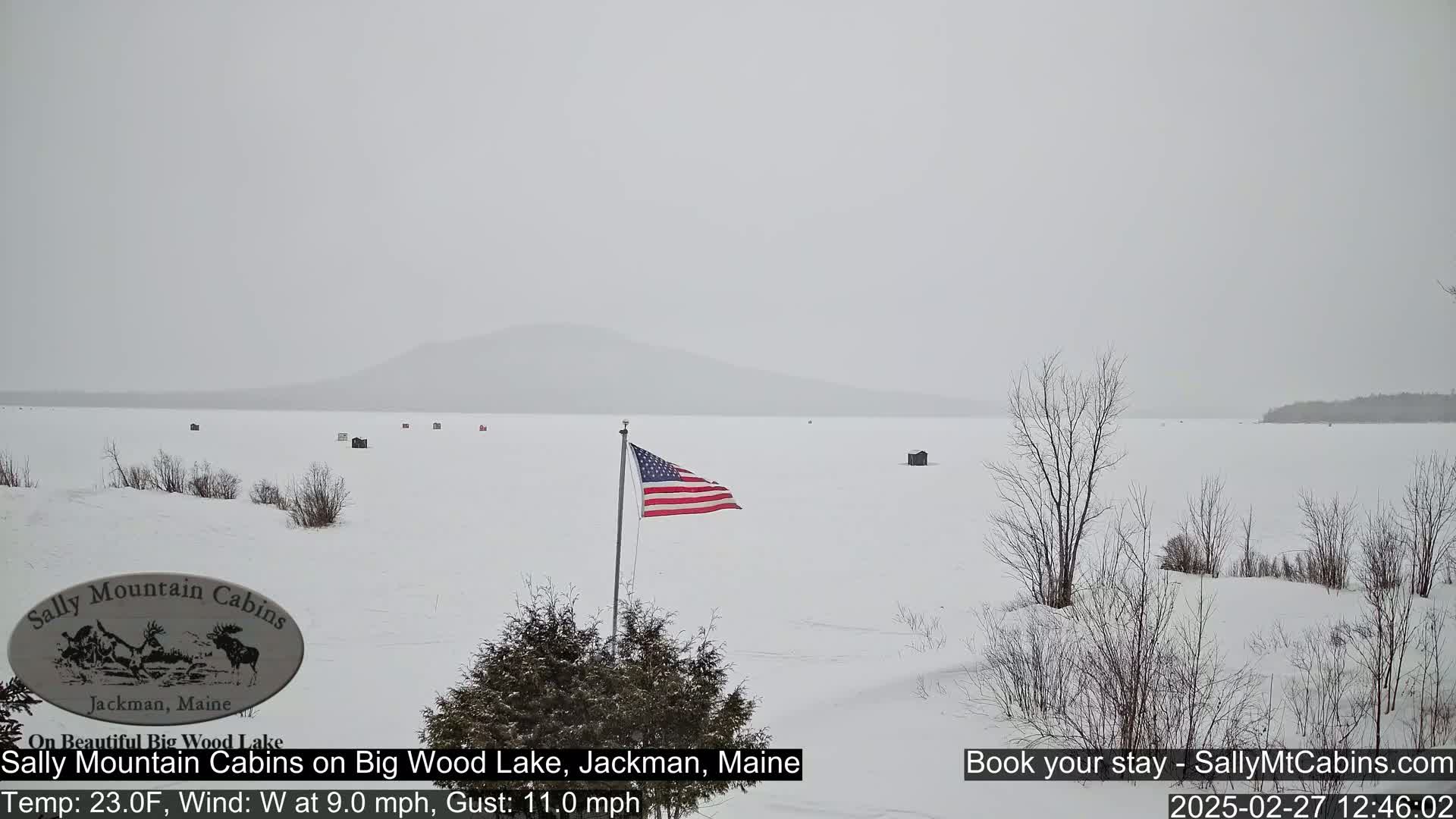 A snow-covered, frozen lake stretches to a distant mountain range under a gray, snowy sky, with a few ice fishing shacks scattered across the ice and an American flag waving in the wind.