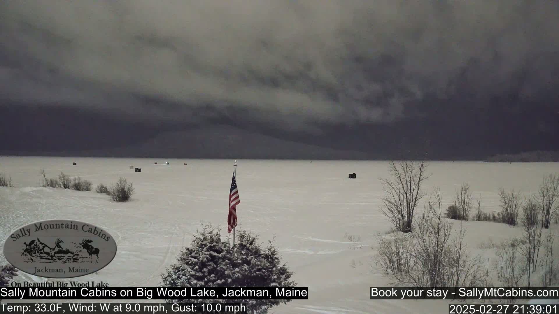 A snow-covered frozen lake with several small structures and a US flag on a pole, under a dark, overcast sky.