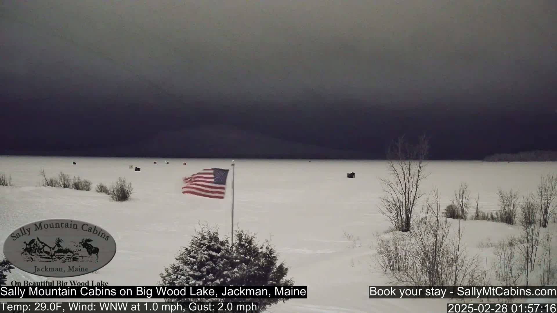 A snow-covered frozen lake is visible under a dark, overcast sky, with a few small structures and an American flag waving in the light wind.