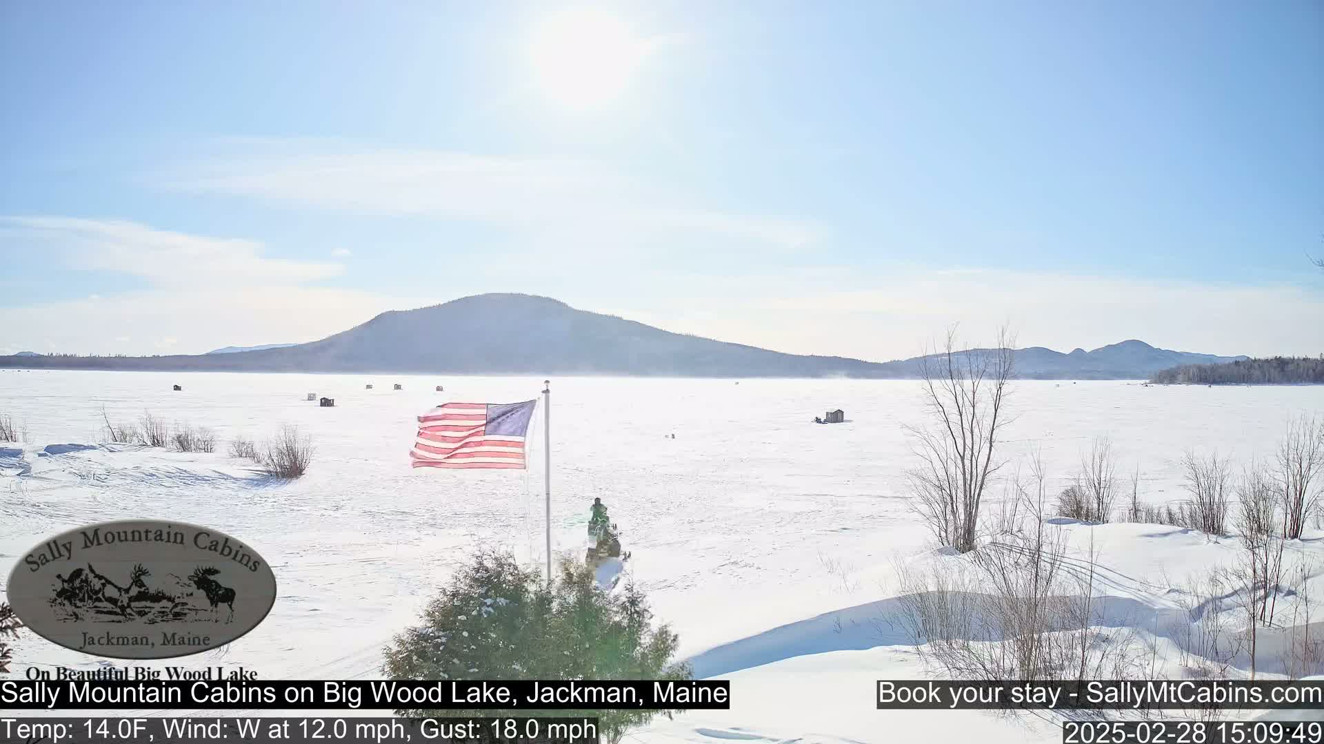 A snow-covered frozen lake with several ice fishing huts, a waving American flag, and snowmobiles is visible under a sunny sky, with mountains in the background.