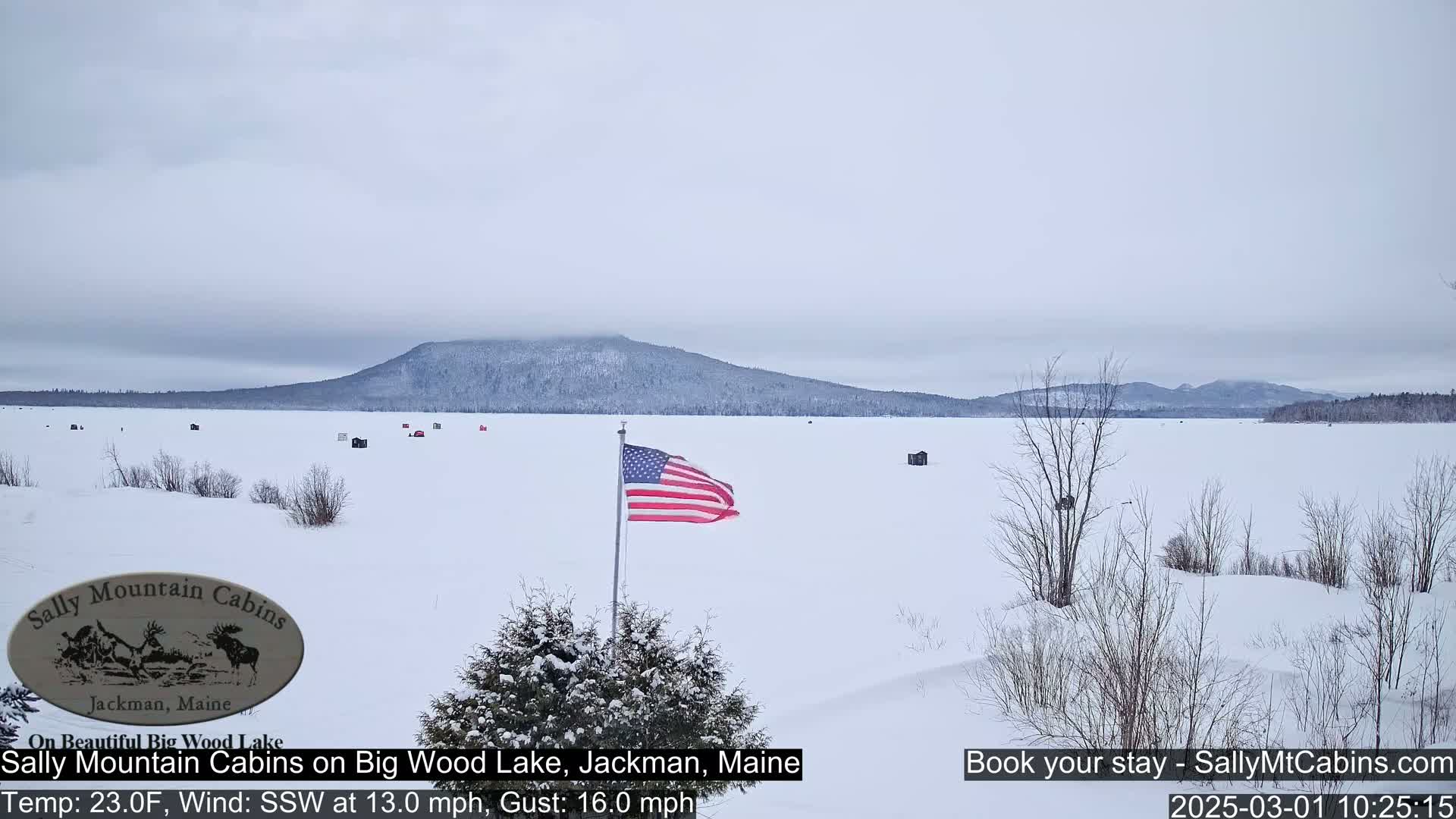 A snow-covered frozen lake stretches to a distant, snow-dusted mountain range under an overcast sky, with a few ice fishing shacks and an American flag visible.
