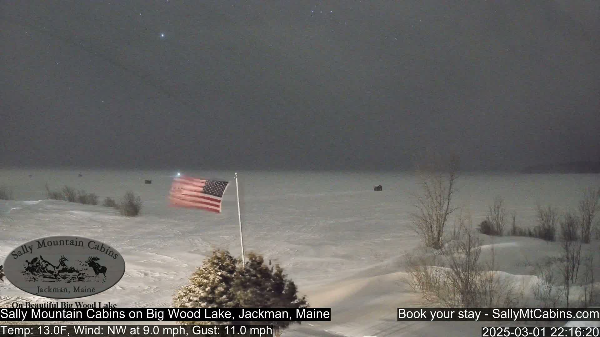 A snow-covered landscape at night shows a frozen lake under a mostly cloudy sky with a few stars visible, an American flag, and some bare trees.