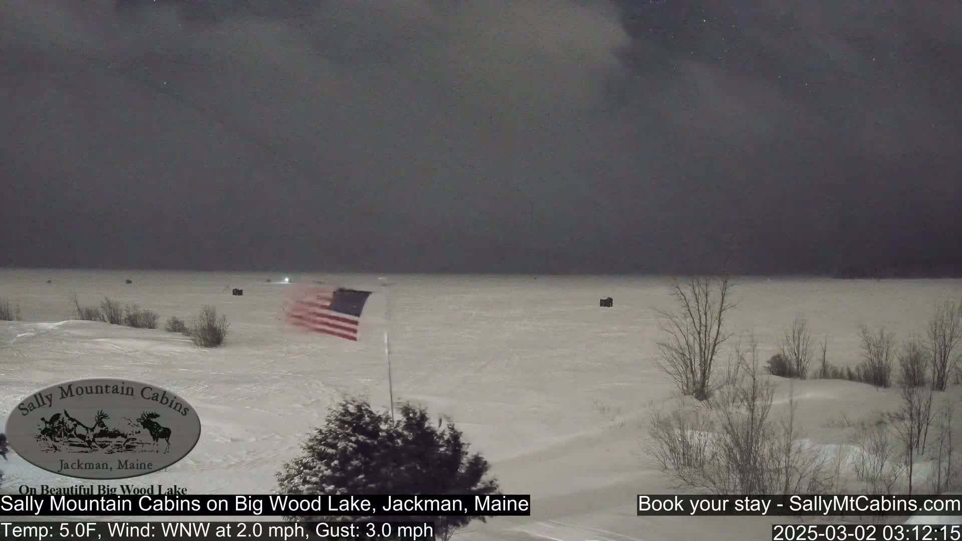 A snow-covered lake is visible under a dark, overcast night sky, with a small American flag waving near the shoreline and some dark structures scattered across the frozen water.