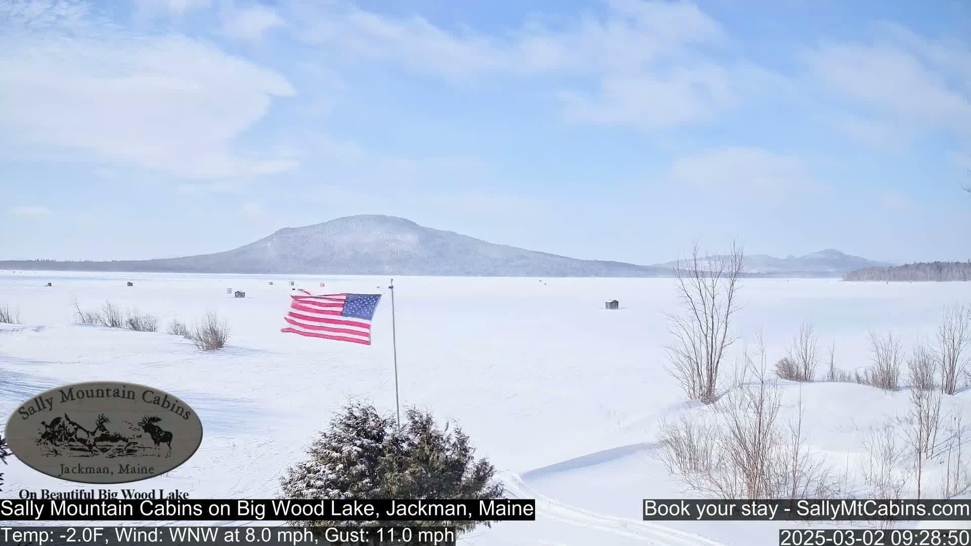 A frozen lake stretches out under a mostly clear sky, with a distant mountain range and an American flag waving in the light wind.