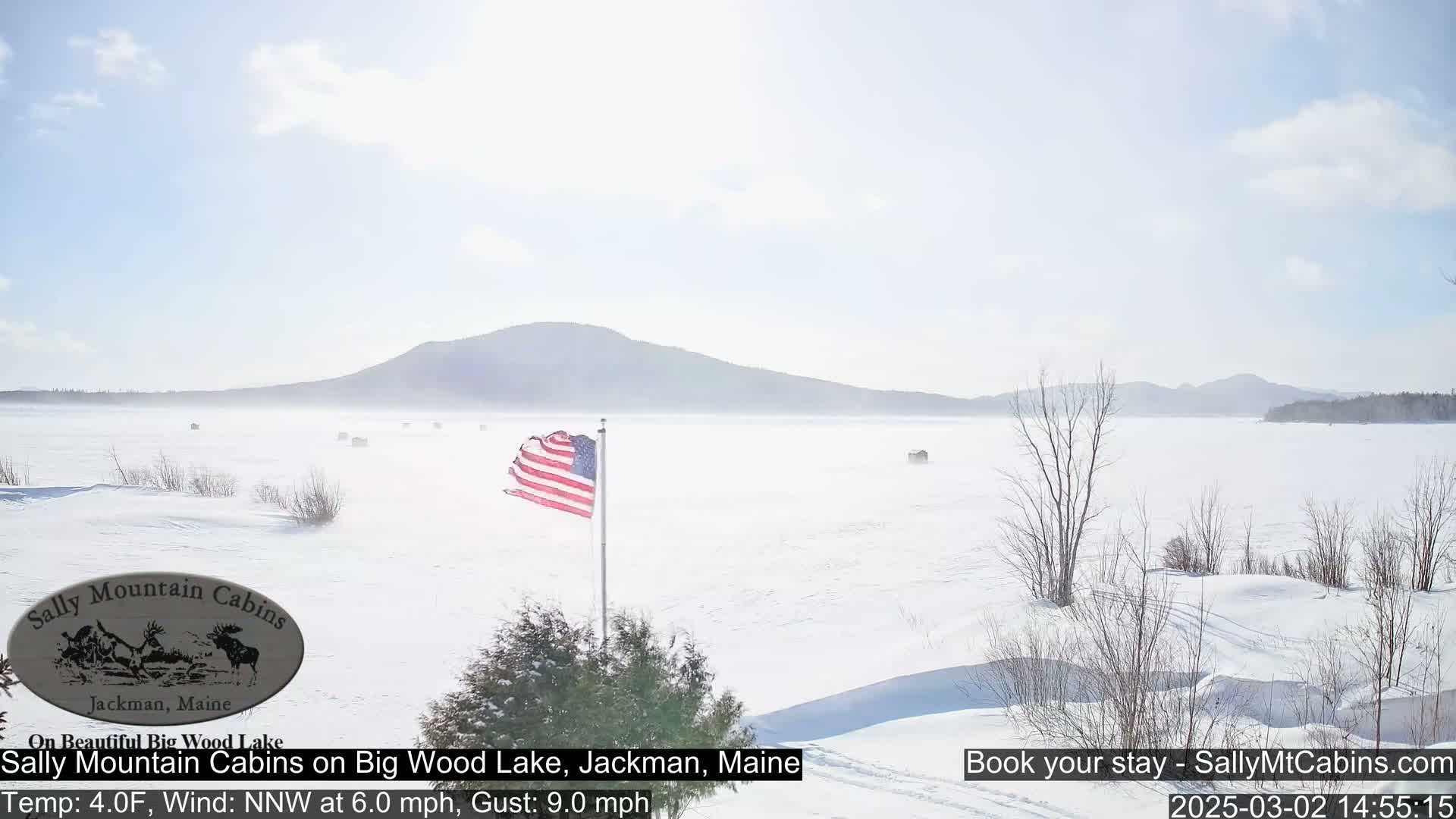 A snow-covered frozen lake stretches to a hazy mountain range under a bright, sunny sky, with an American flag waving in the wind.