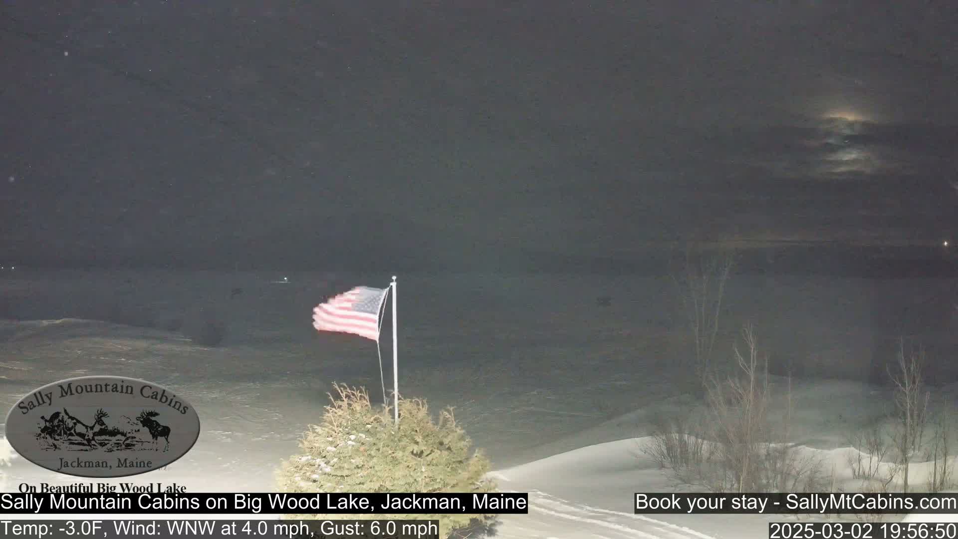 A nighttime view of a snow-covered landscape with a frozen lake, an American flag waving in a WNW wind, and sparsely-leafed shrubs in the foreground under a mostly cloudy sky.