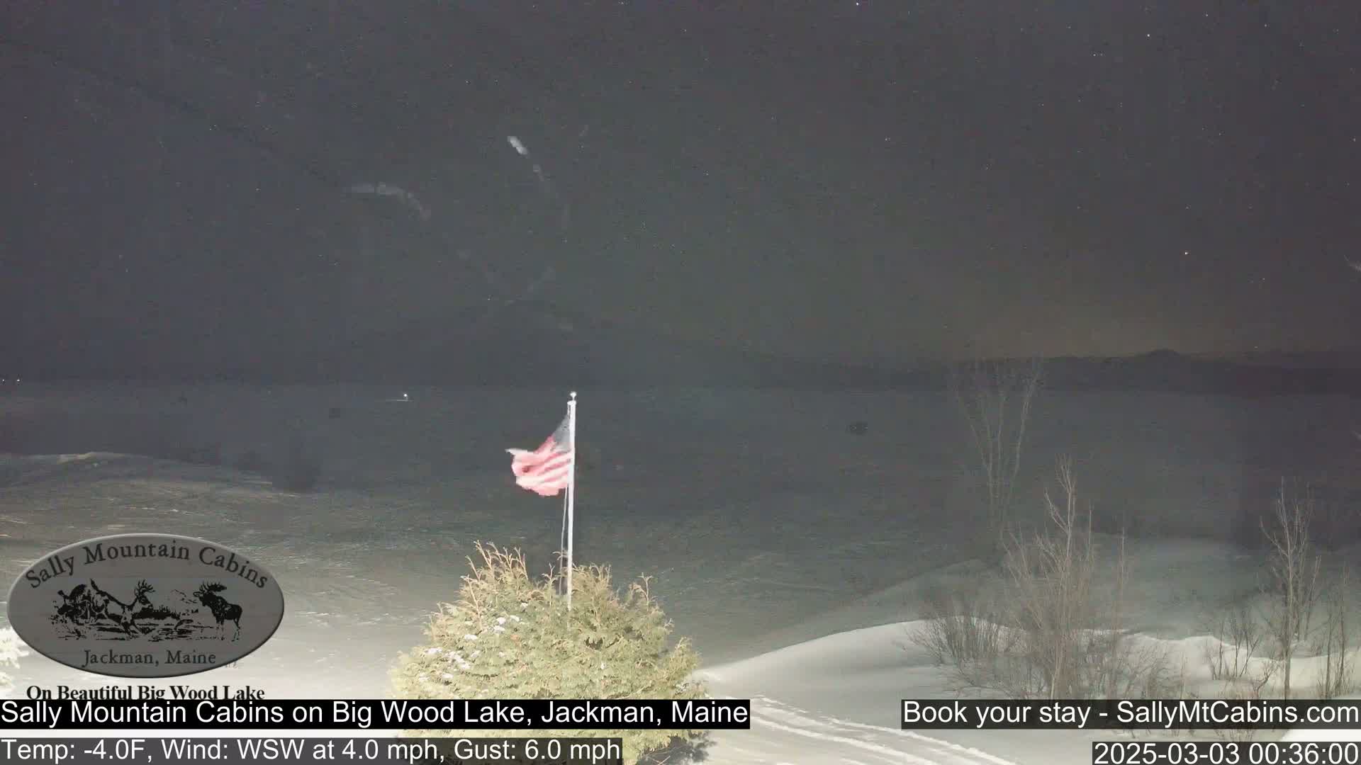 A nighttime view of a snow-covered landscape with a frozen lake and an American flag waving in a 4 mph wind with 6 mph gusts, under a mostly clear, starry sky.