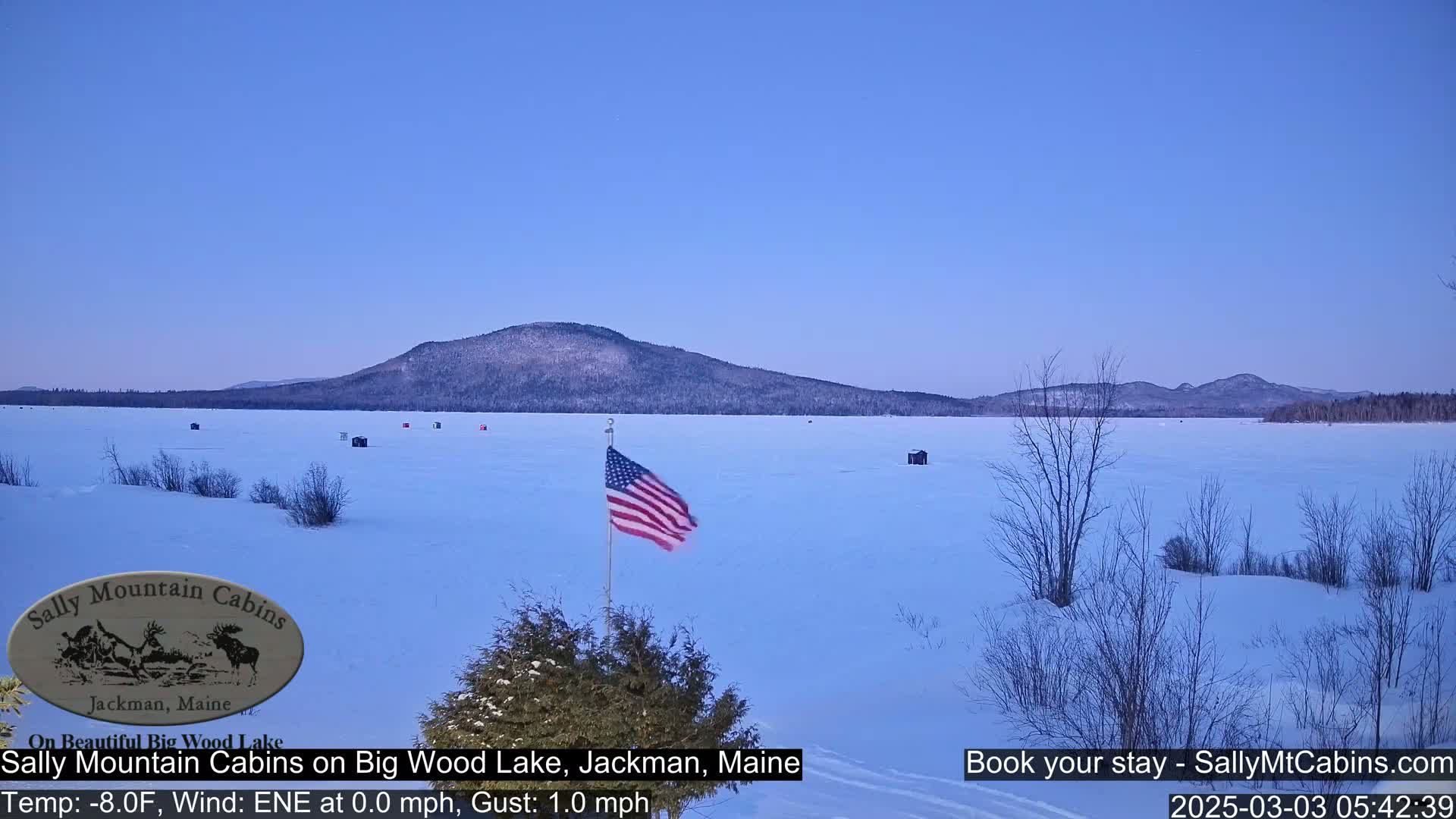 A frozen lake under a clear, twilight sky, with a few ice fishing shacks and an American flag in the foreground, and a snow-covered mountain in the distance.