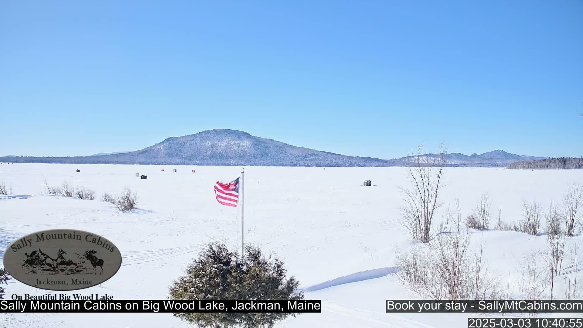 A snow-covered frozen lake stretches to a distant mountain range under a clear blue sky, with a few small ice fishing huts and an American flag visible.