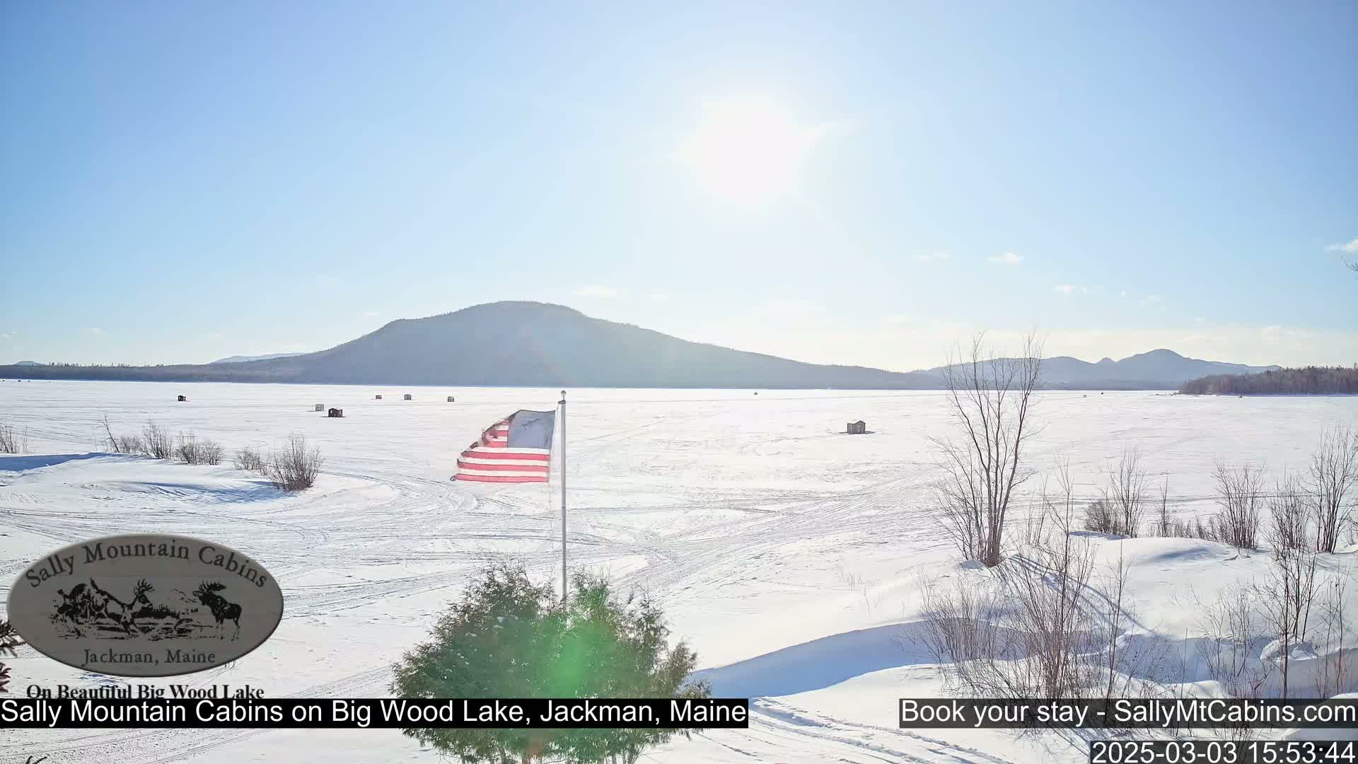 A frozen lake stretches out under a sunny sky, with a few ice fishing huts scattered across its surface and a distant mountain range visible in the background.