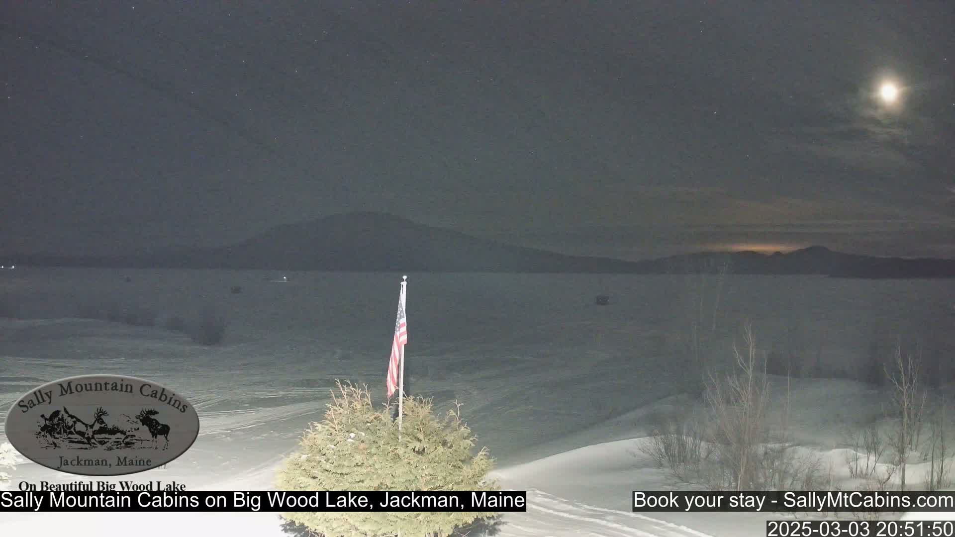 A nighttime view of a snow-covered lake and distant mountains under a partly cloudy sky with a bright moon.