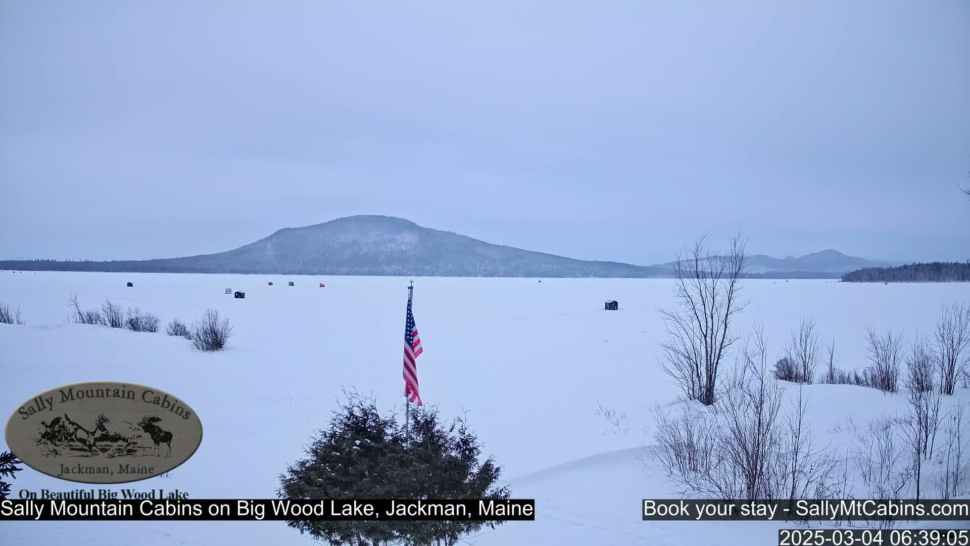 A snow-covered frozen lake stretches to a distant mountain range under a cloudy, pale sky, with several small ice fishing huts scattered across the ice and an American flag displayed in the foreground.
