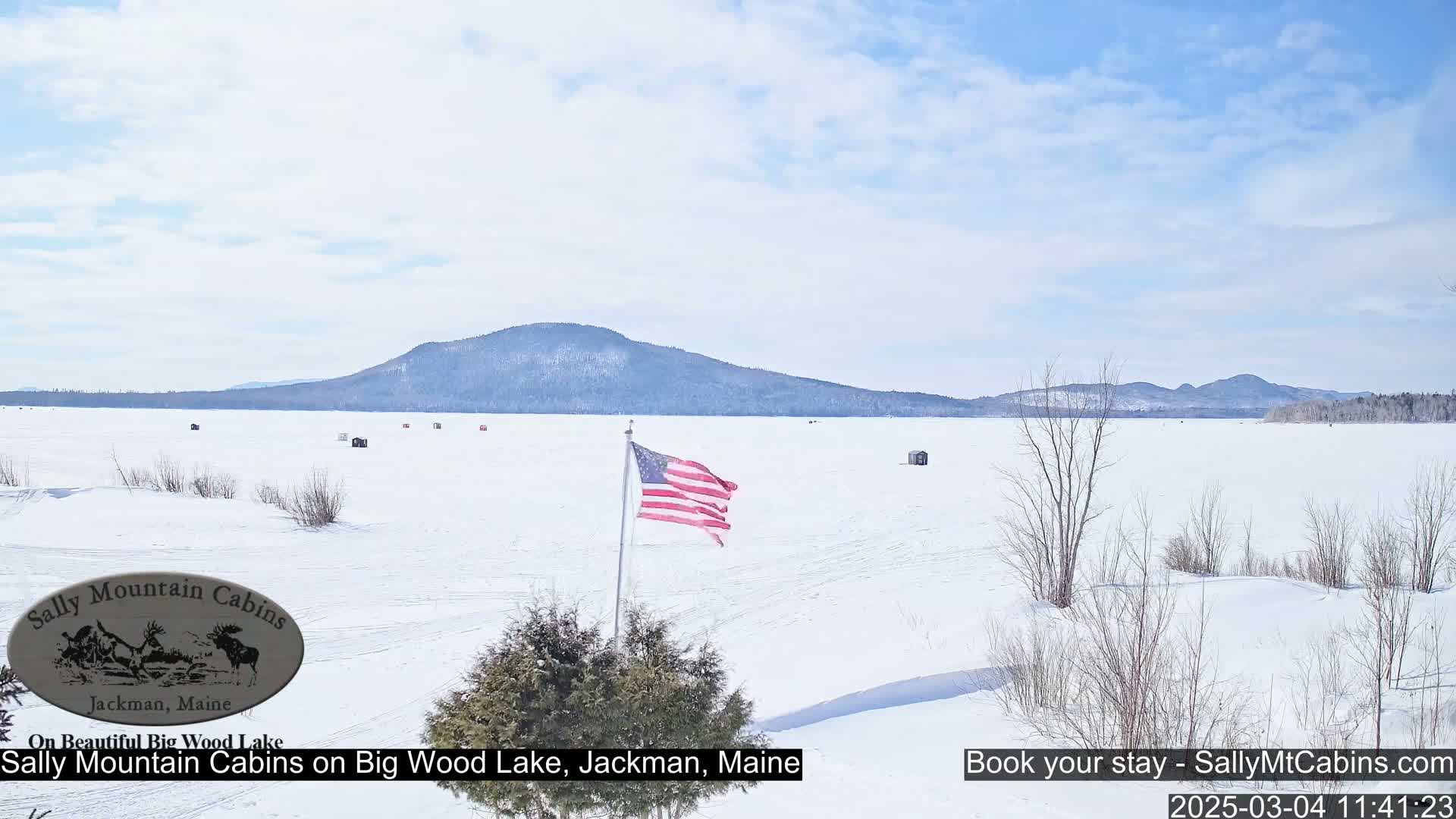 A frozen lake stretches to a snow-covered mountain range under a mostly clear sky, with a few ice fishing shelters scattered across the ice and an American flag waving near the foreground.