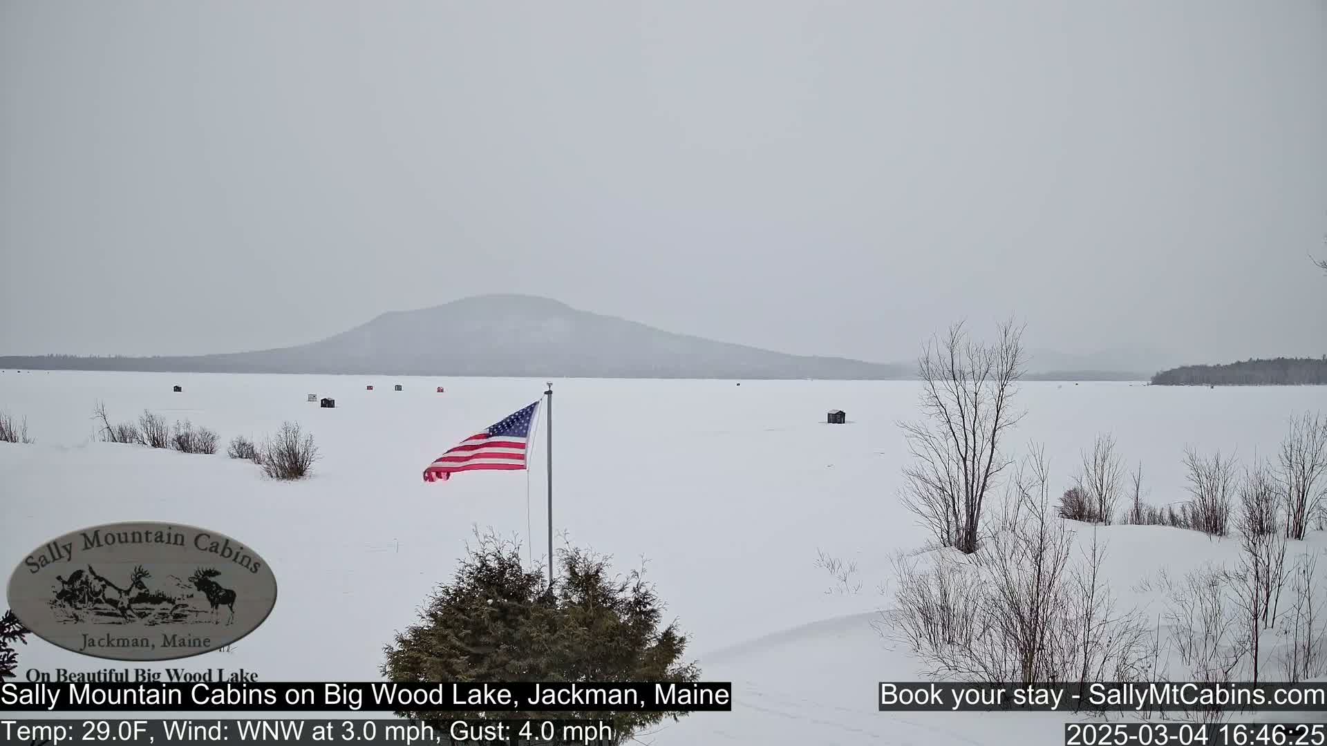 A snow-covered frozen lake stretches to a distant, hazy mountain under an overcast sky, with a few ice fishing huts and an American flag visible.