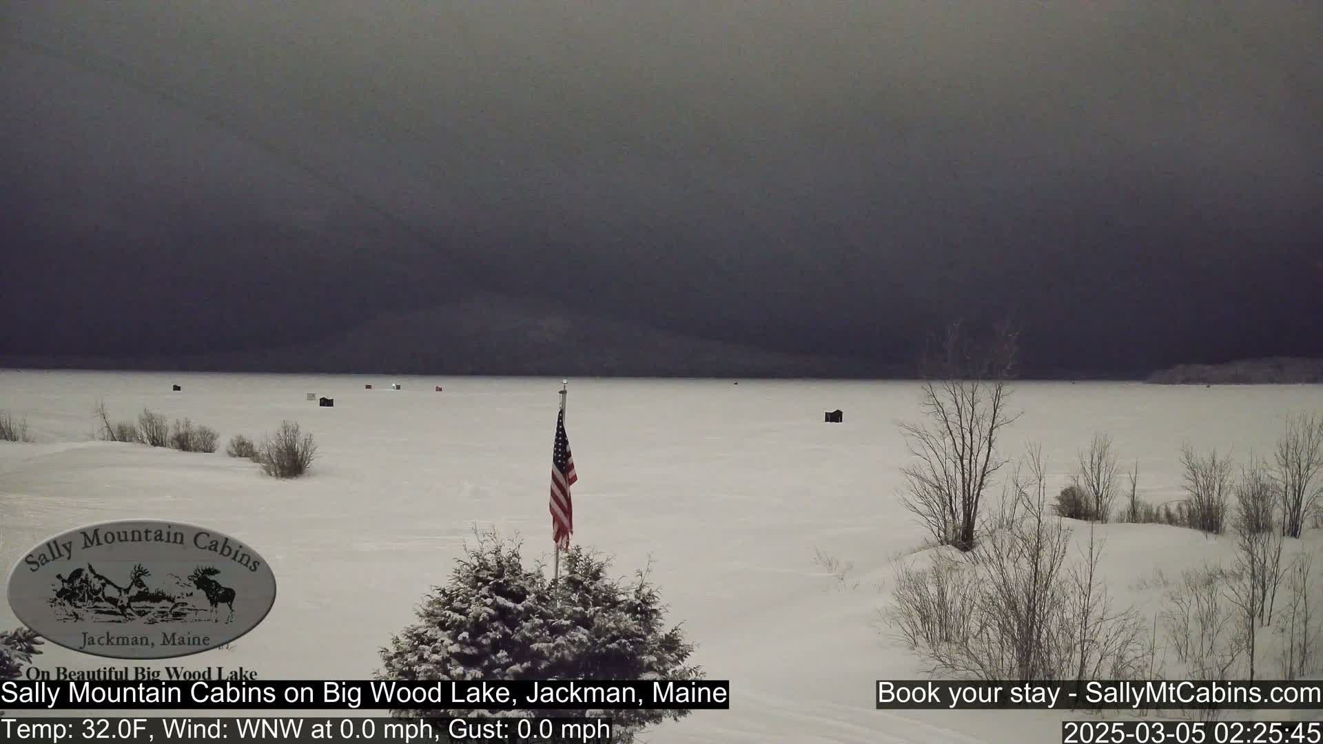 A snow-covered frozen lake is visible under a dark, overcast sky, with a few ice fishing shelters scattered across the ice and an American flag planted in the snow near the foreground.