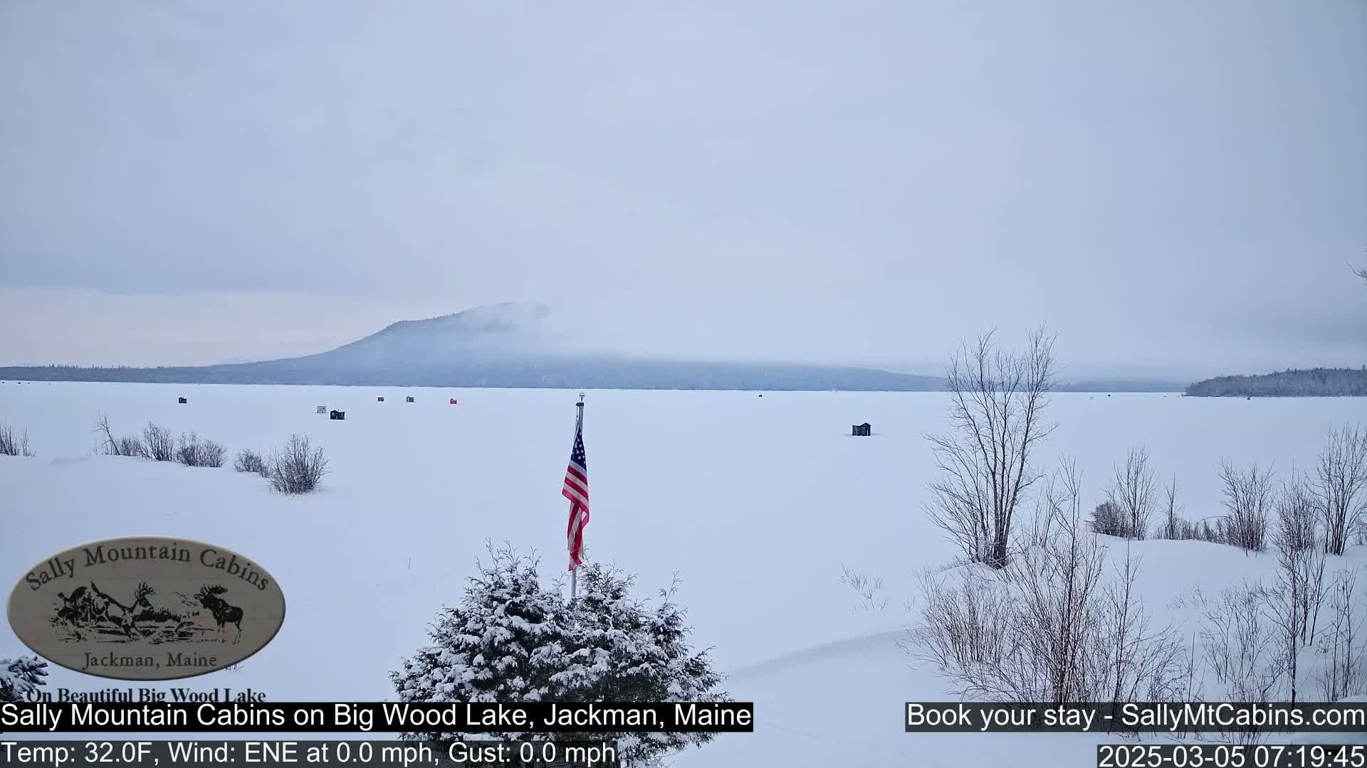 A snow-covered frozen lake stretches to a hazy mountain range under a gray sky, with a few ice fishing shacks scattered across the ice and an American flag planted near the foreground.