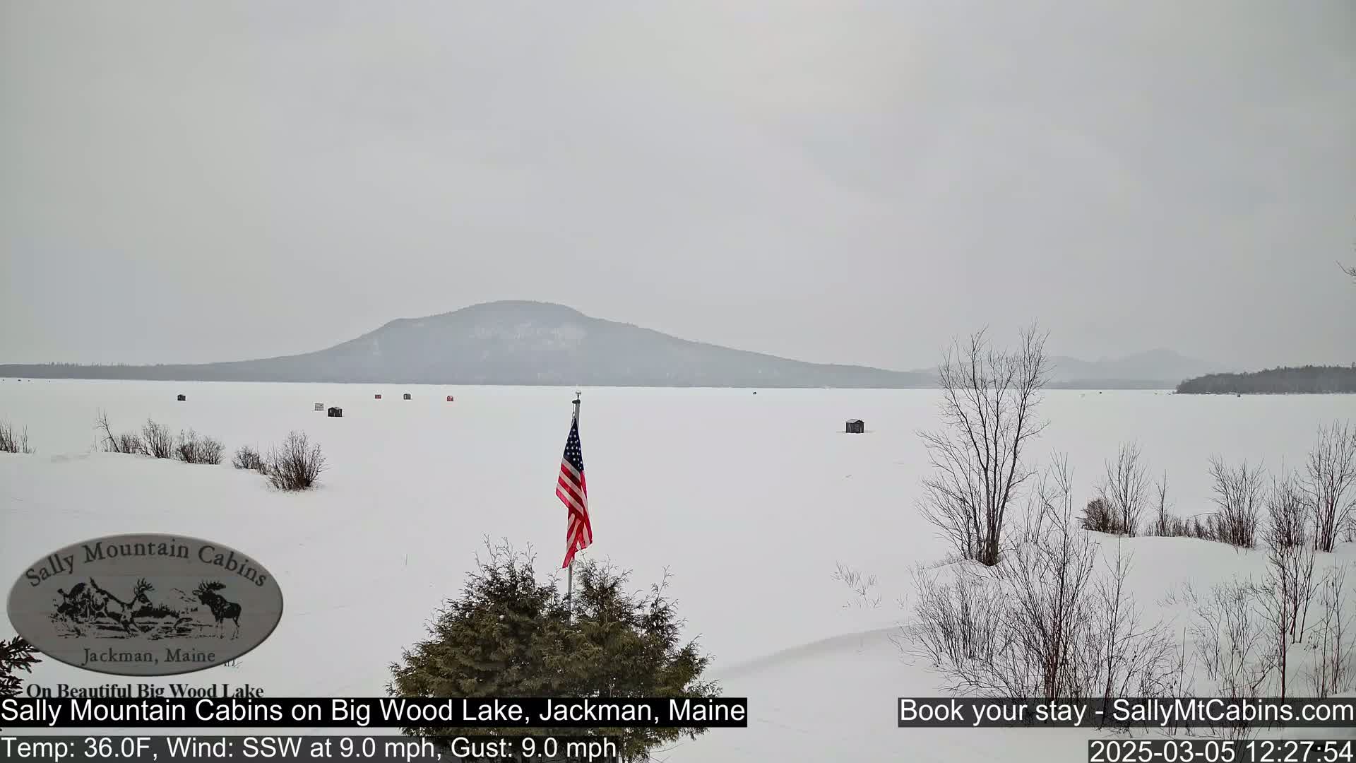 A snow-covered frozen lake stretches to a distant mountain range under an overcast sky, with a few ice fishing huts visible and an American flag planted in the foreground.