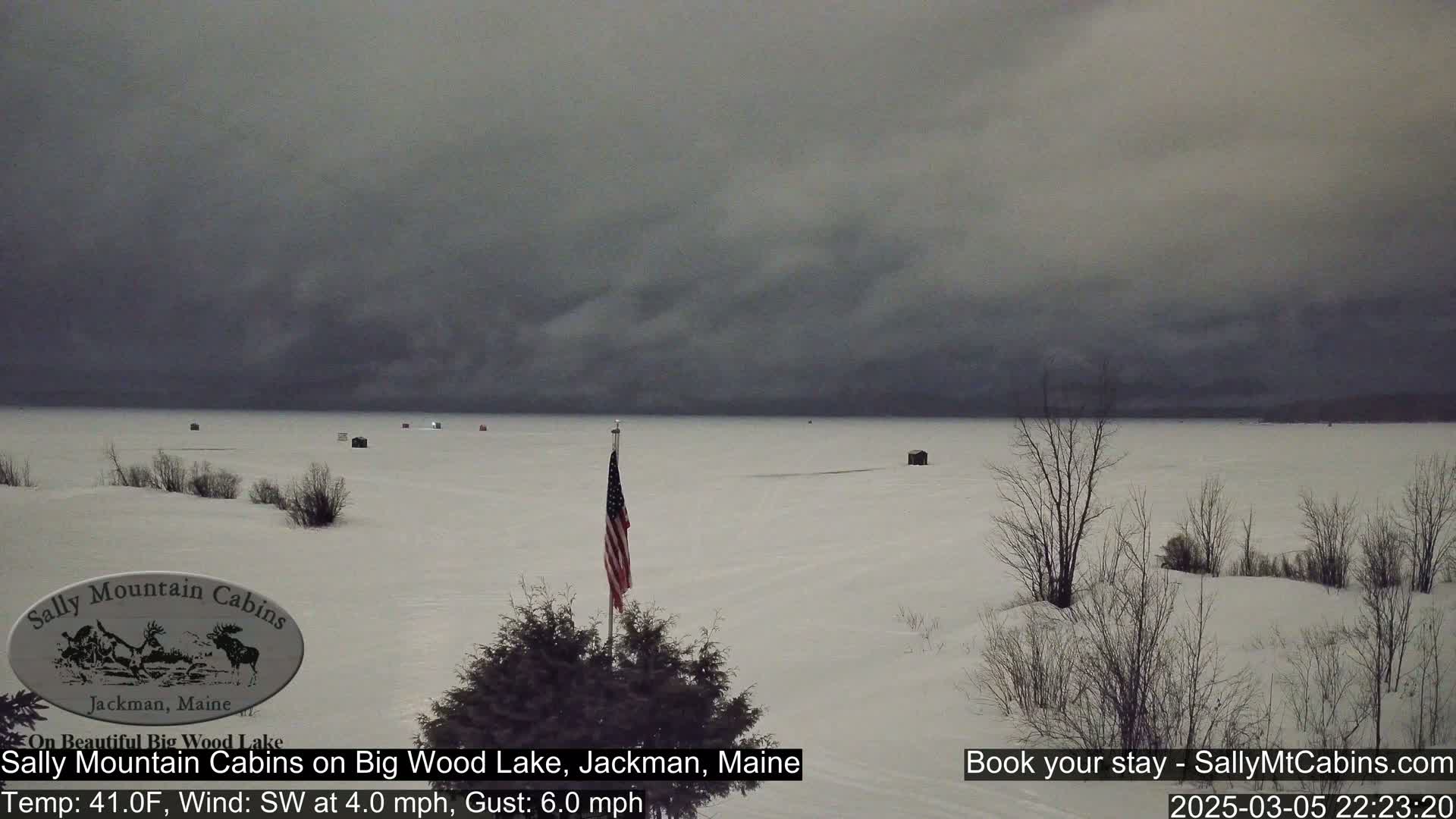 A snow-covered frozen lake with several small structures and a lone American flag, under an overcast sky.