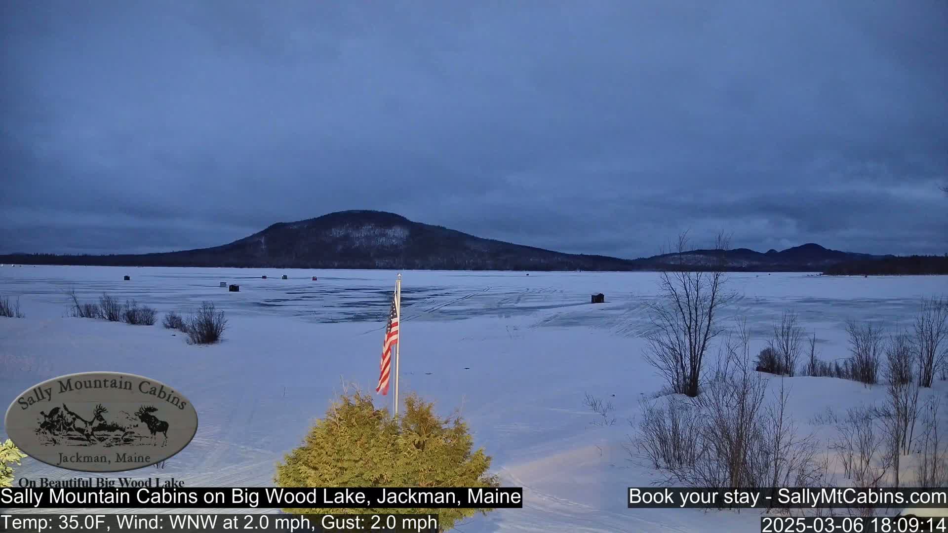A frozen lake under a twilight sky, with a few ice fishing shacks and an American flag visible, is backed by a dark mountain range.
