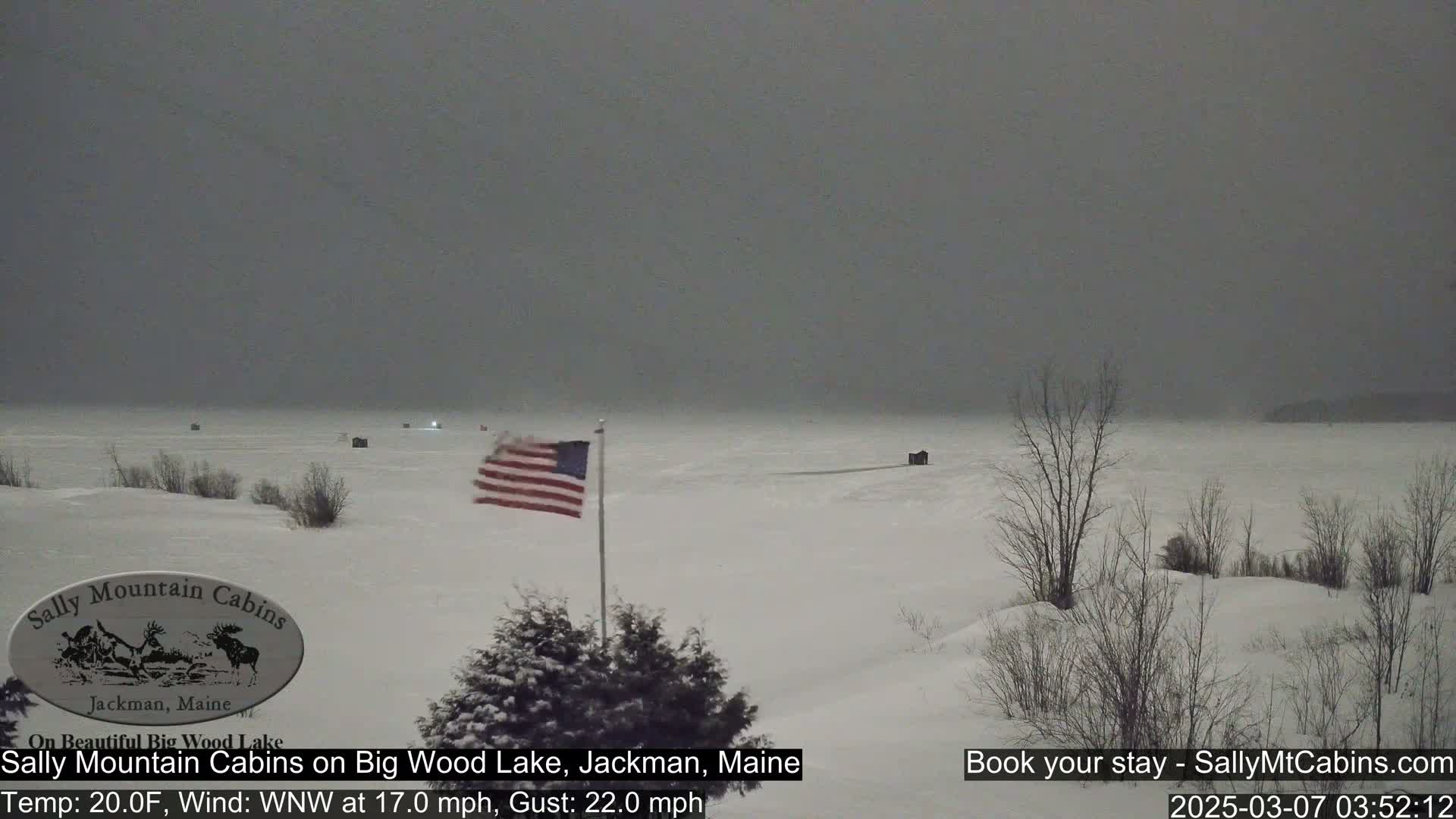 A snow-covered frozen lake is seen under a gray, overcast sky, with a few ice fishing huts visible in the distance and an American flag waving near the foreground.