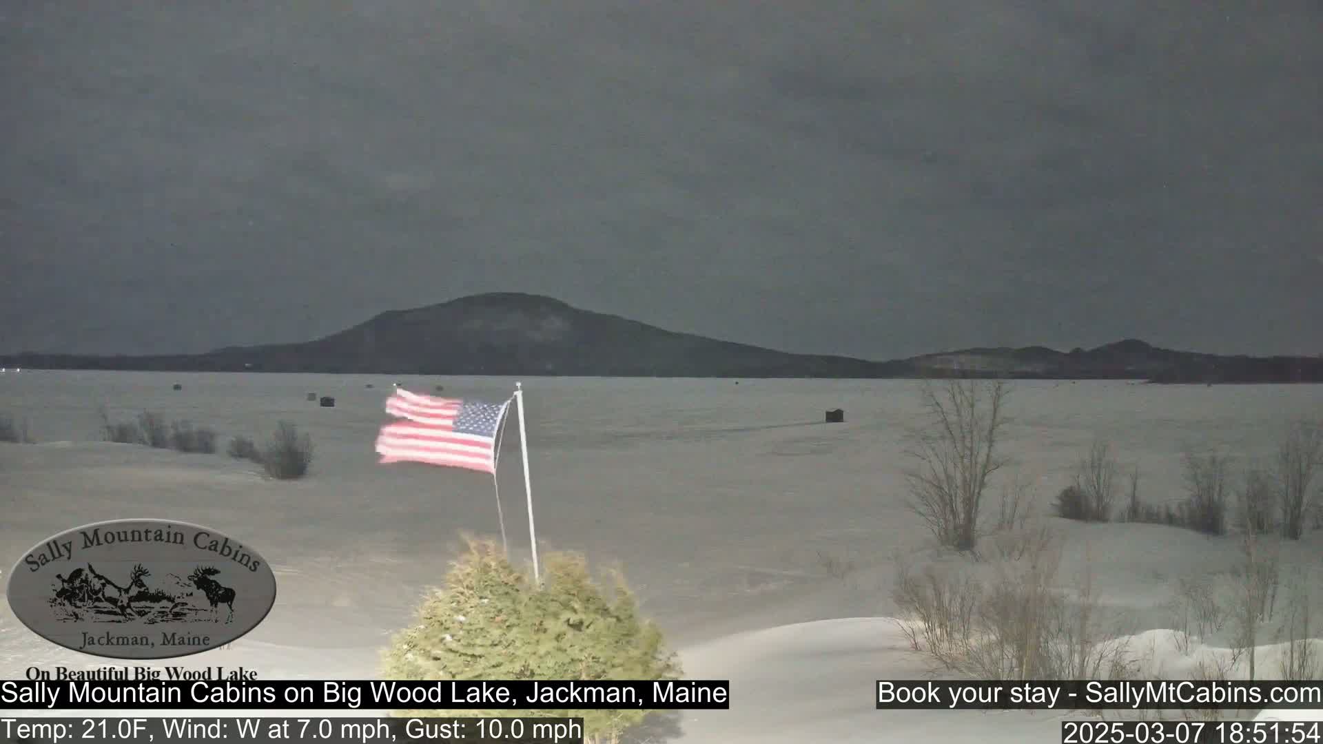 A snow-covered landscape stretches to a frozen lake under a dark, overcast sky, with a small American flag waving in the foreground.