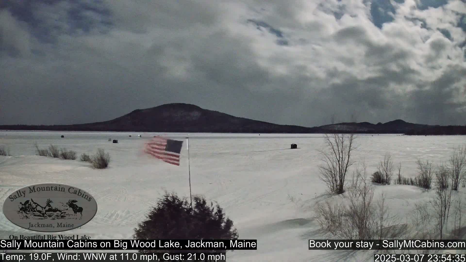 A snow-covered frozen lake stretches out before a dark mountain range under a cloudy sky, with a small American flag planted near the shoreline.