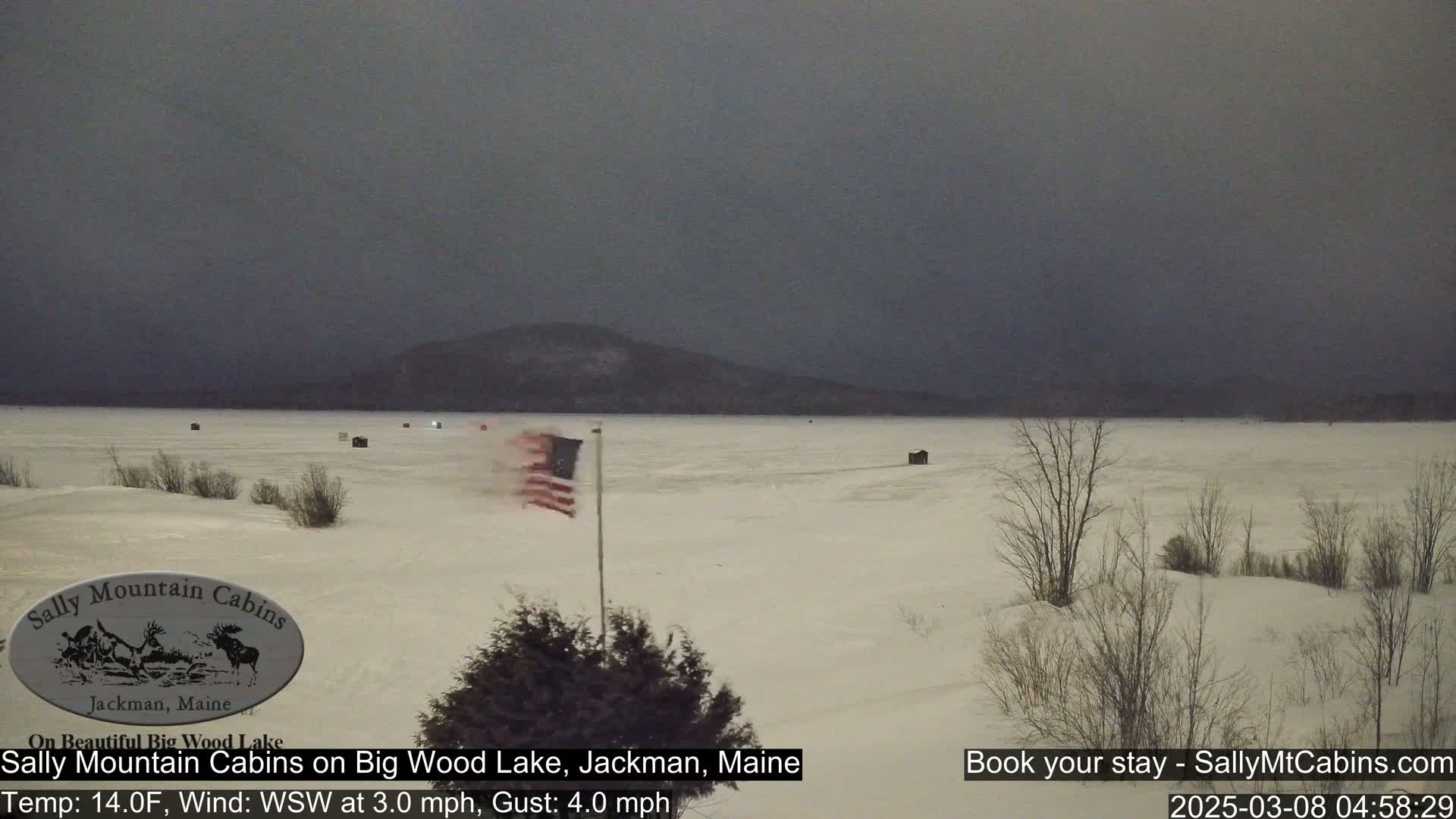 A snow-covered frozen lake is seen under a dark, overcast sky with a few ice fishing shacks visible and a flag waving in the wind.