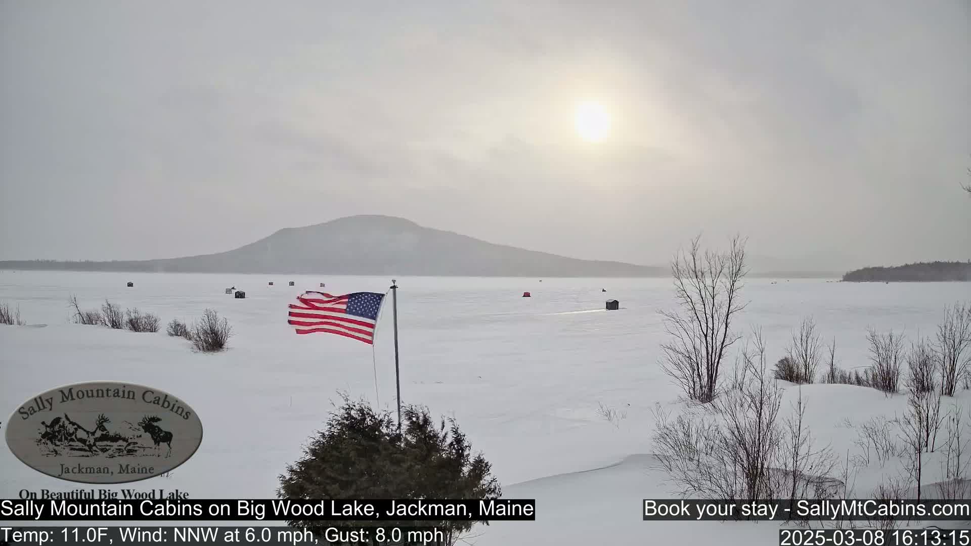 A snow-covered frozen lake stretches to a distant mountain under a hazy, sunlit sky, with a few ice fishing huts scattered across the ice and an American flag waving near the shore.