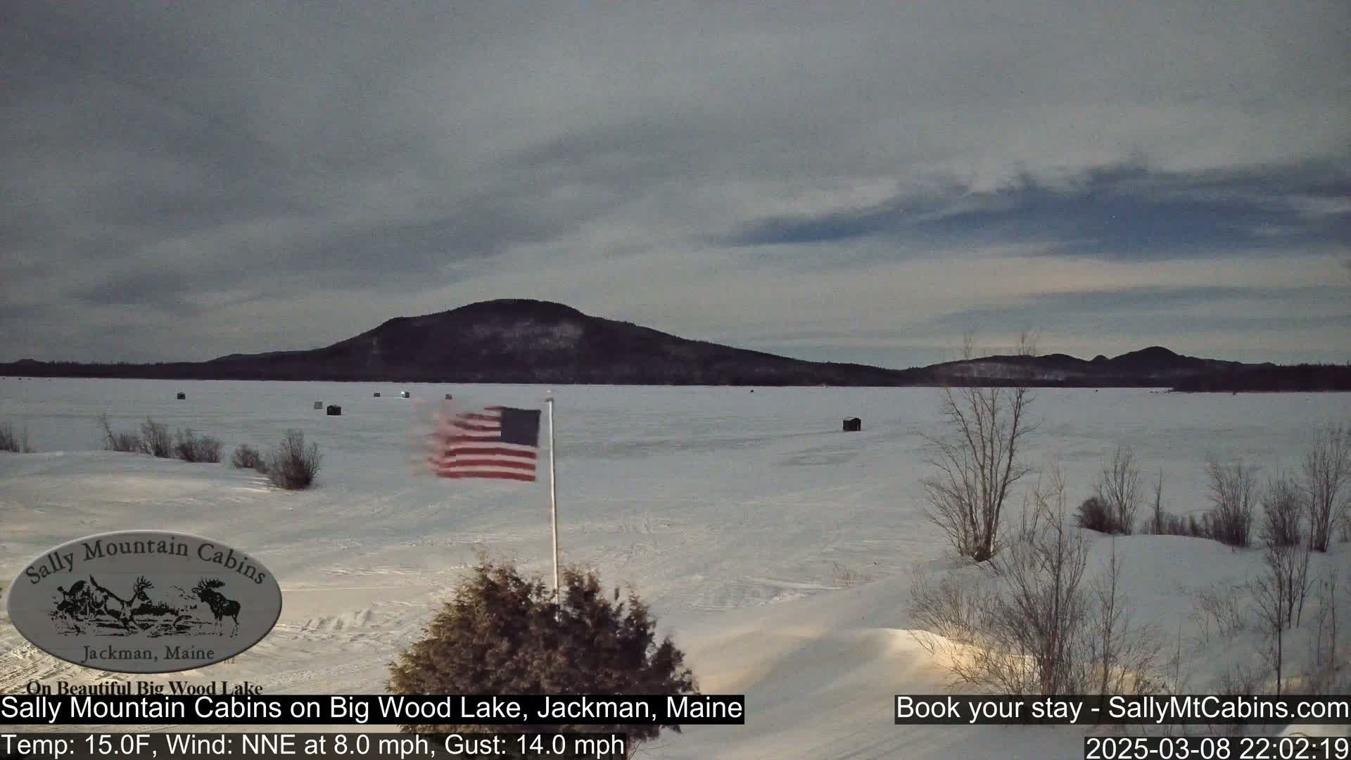 A snow-covered frozen lake stretches out before a distant mountain range under an overcast sky, with an American flag waving near the foreground.