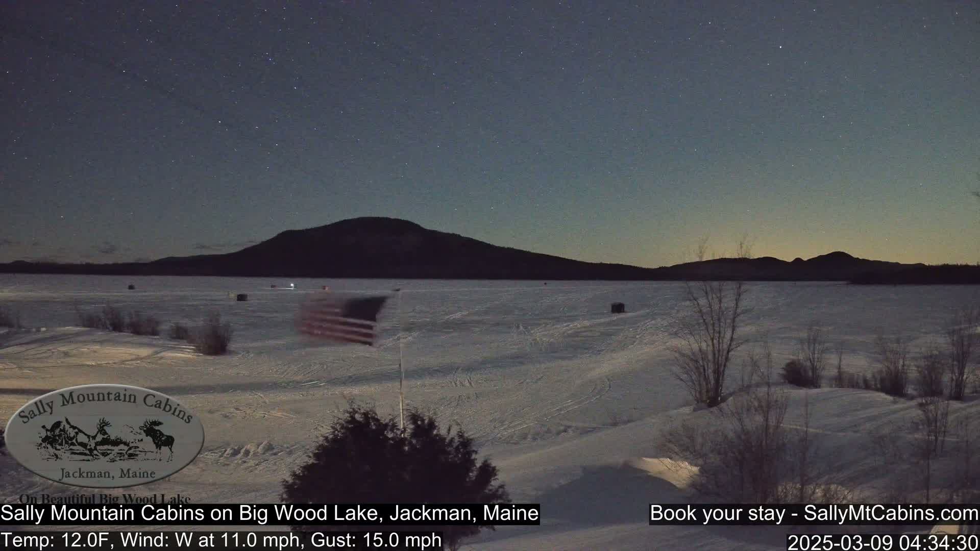 A frozen lake under a starry night sky is viewed from a snow-covered shoreline, with a distant mountain range and an American flag visible.