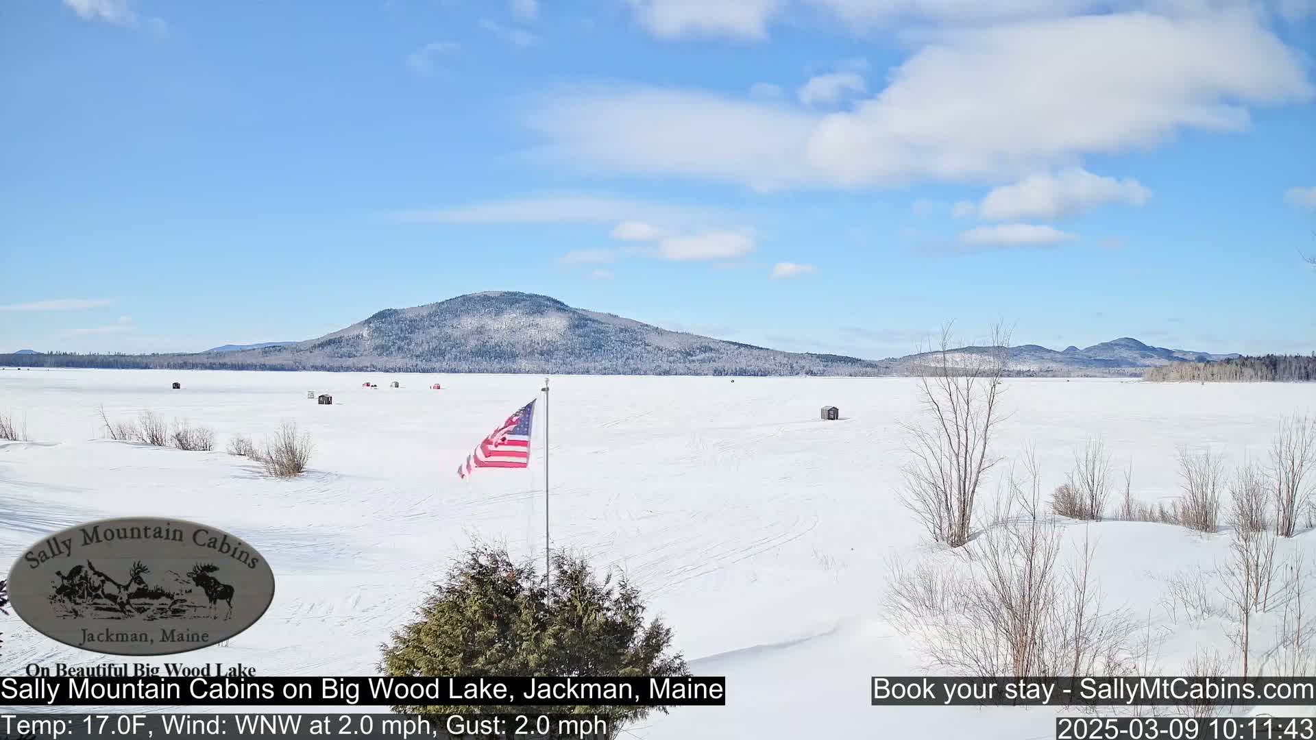 A snow-covered frozen lake stretches to a snow-dusted mountain range under a mostly clear, blue sky.