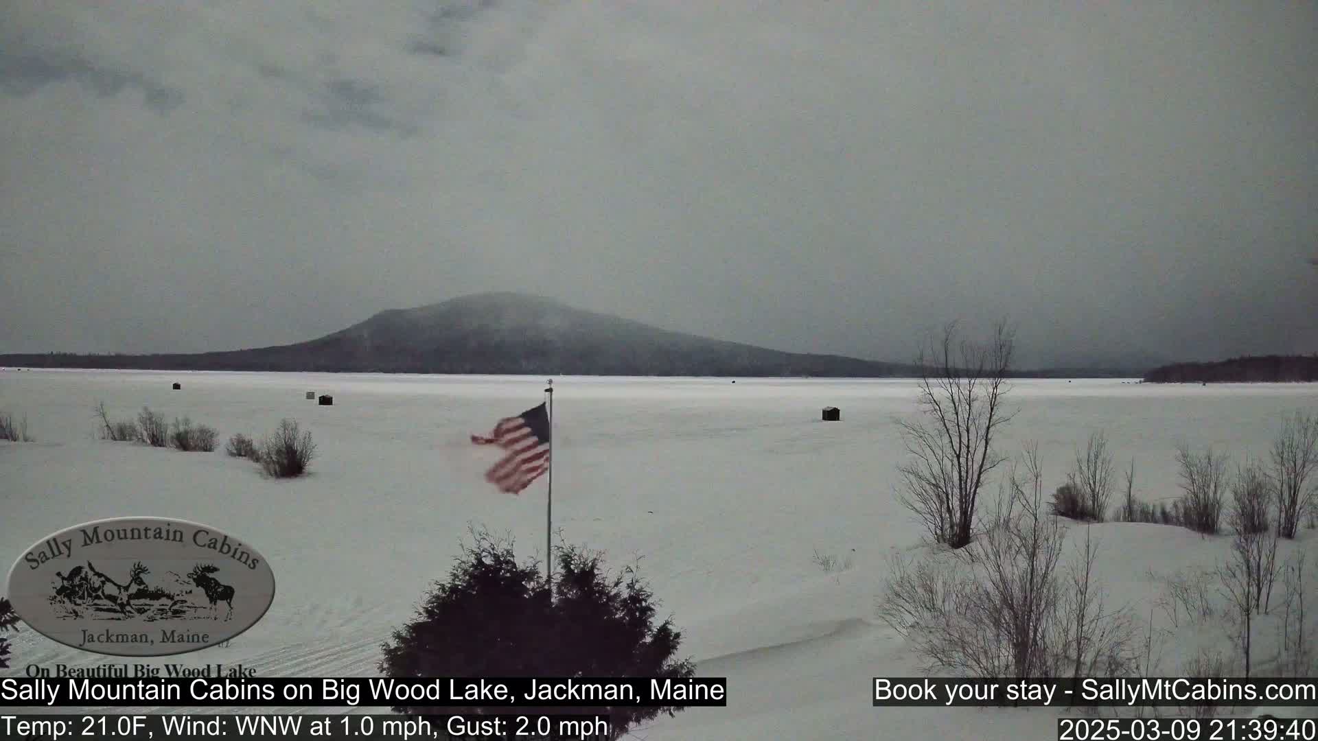 A snow-covered frozen lake stretches to a snow-dusted mountain under an overcast sky, with an American flag waving in the light wind.