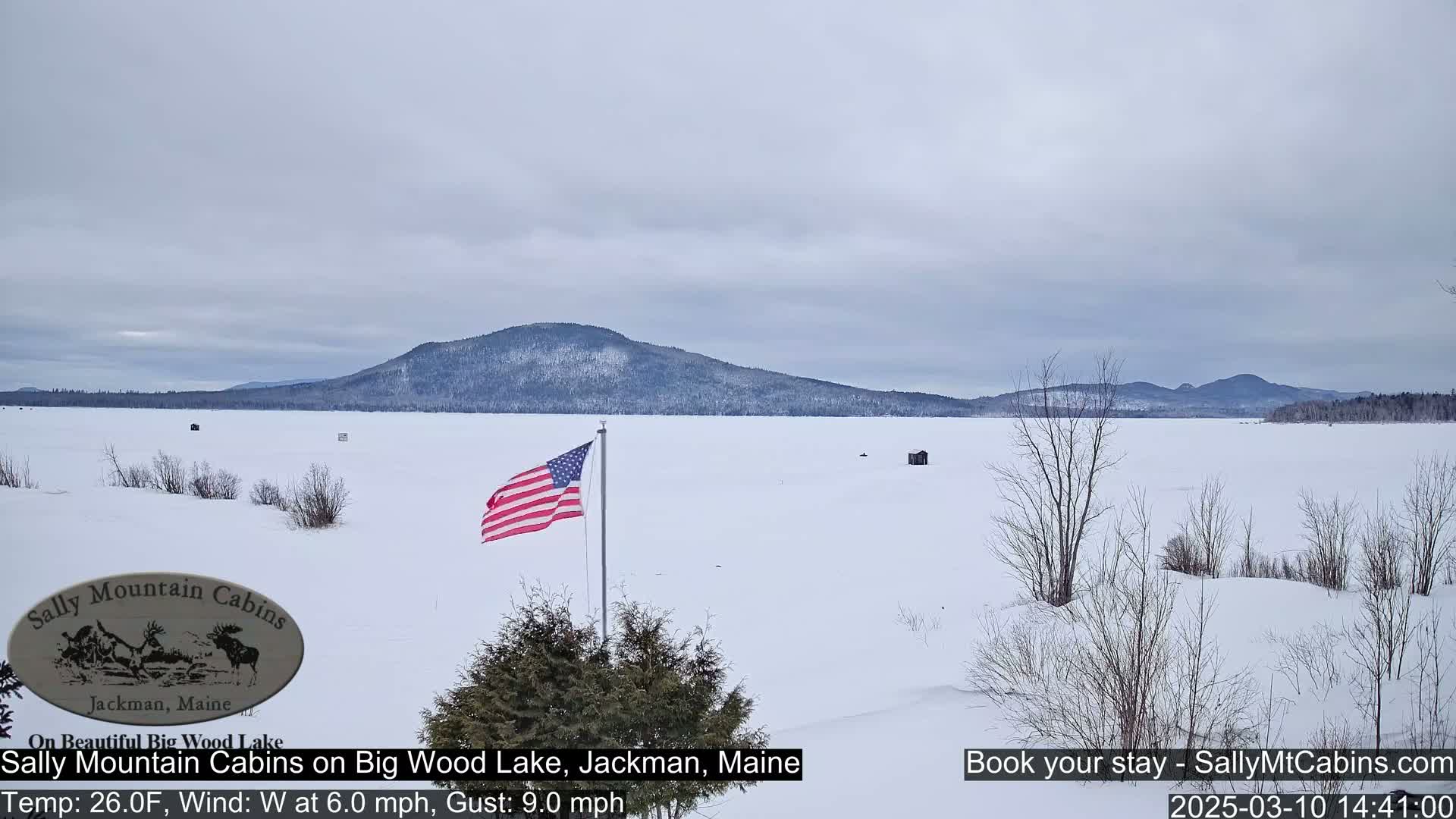 A snow-covered frozen lake stretches to a distant mountain range under a gray sky, with an American flag in the foreground.