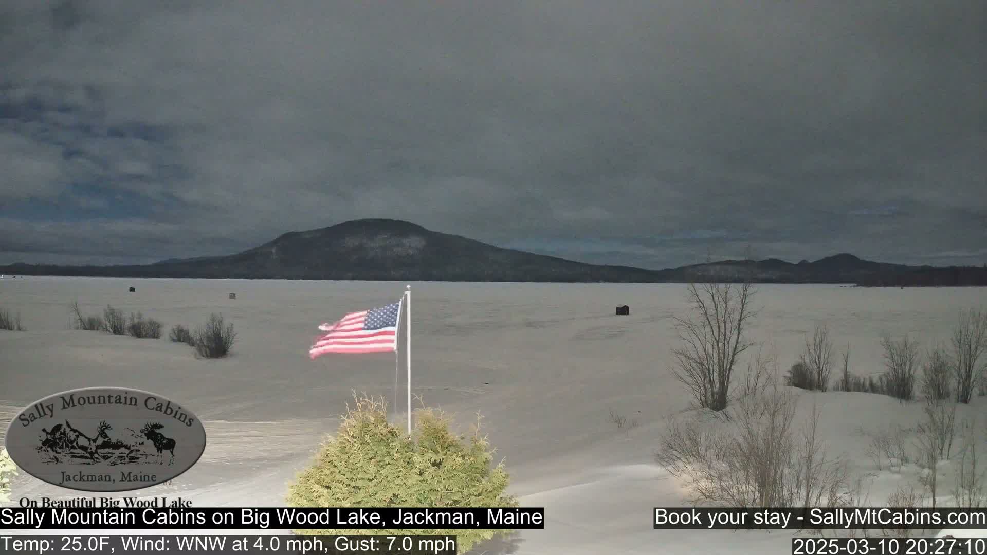 A snow-covered frozen lake stretches out to a dark, cloudy sky, under which a distant mountain range is visible; an American flag waves from a pole in the foreground.