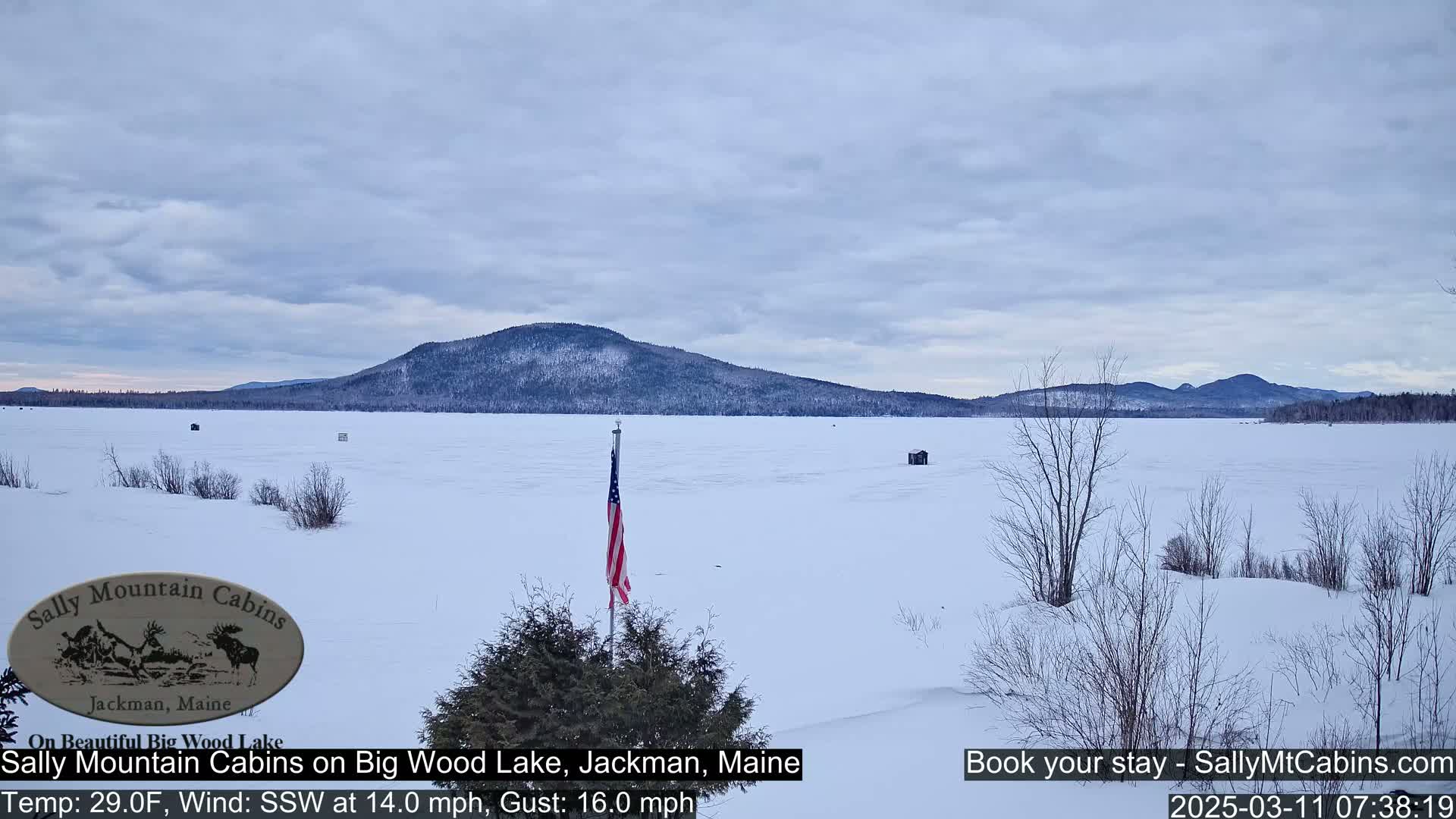 A snow-covered frozen lake stretches to a wooded mountain range under a mostly cloudy sky.