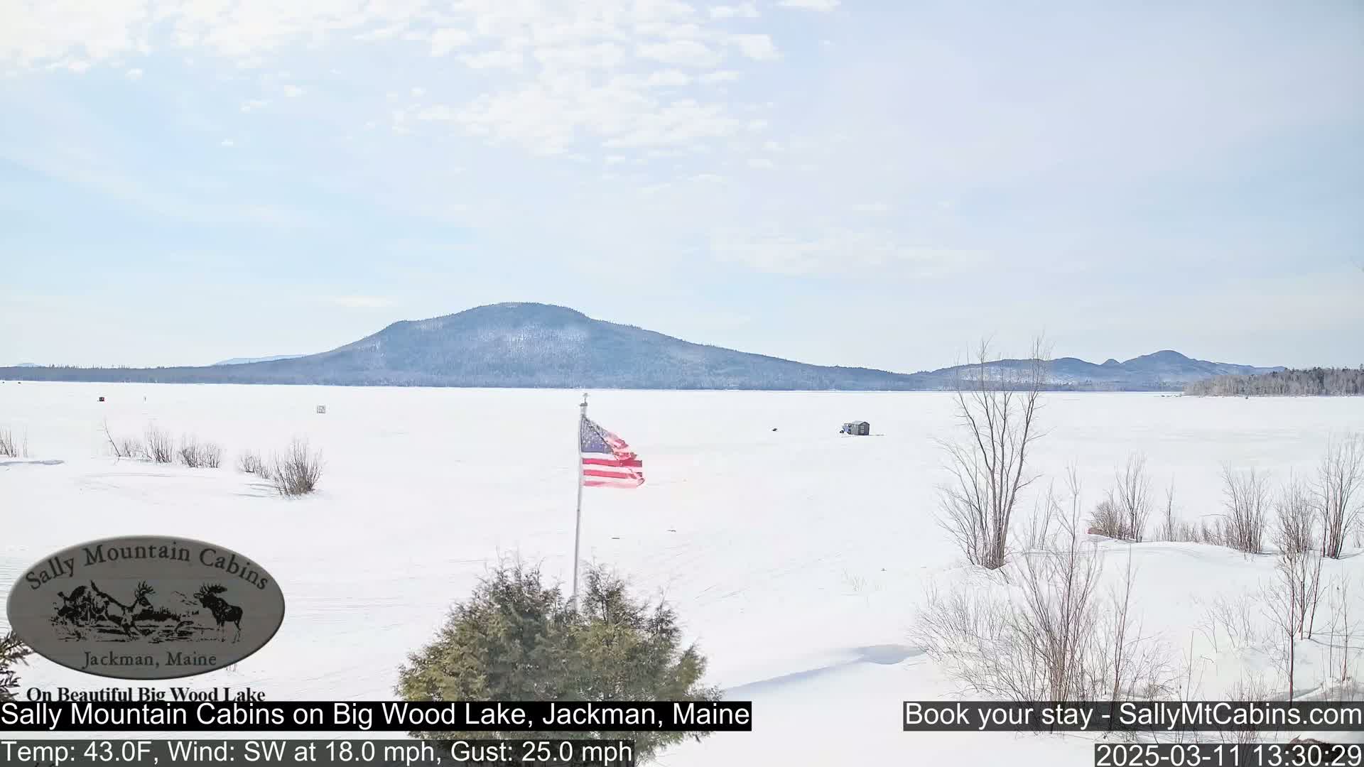 A snow-covered frozen lake stretches to a distant mountain range under a partly cloudy sky, with a small American flag planted near the foreground.