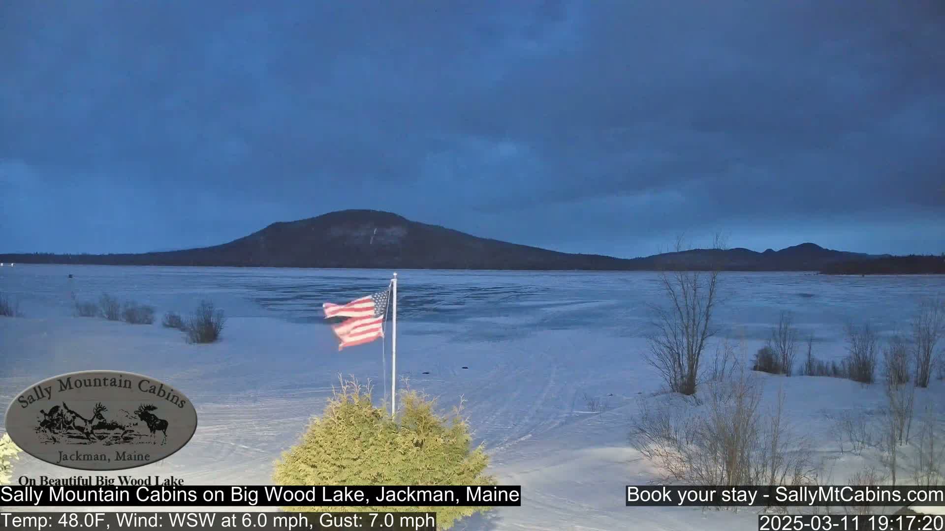 A partly frozen lake stretches out before a snow-covered shoreline under a twilight sky, with an American flag waving in a light wind.