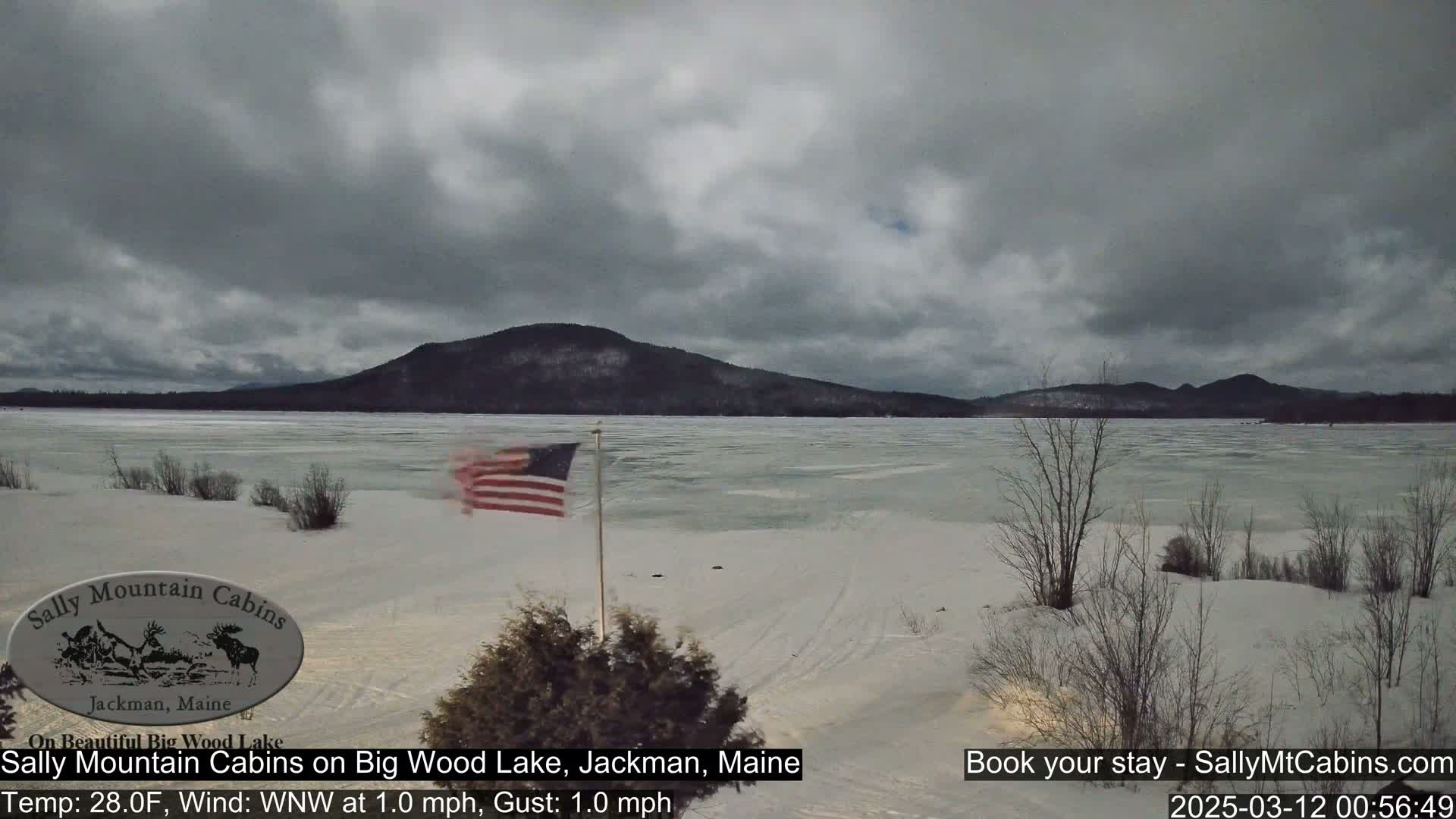 A partially frozen lake stretches toward a snow-dusted mountain range under a cloudy sky, with an American flag waving in the foreground.