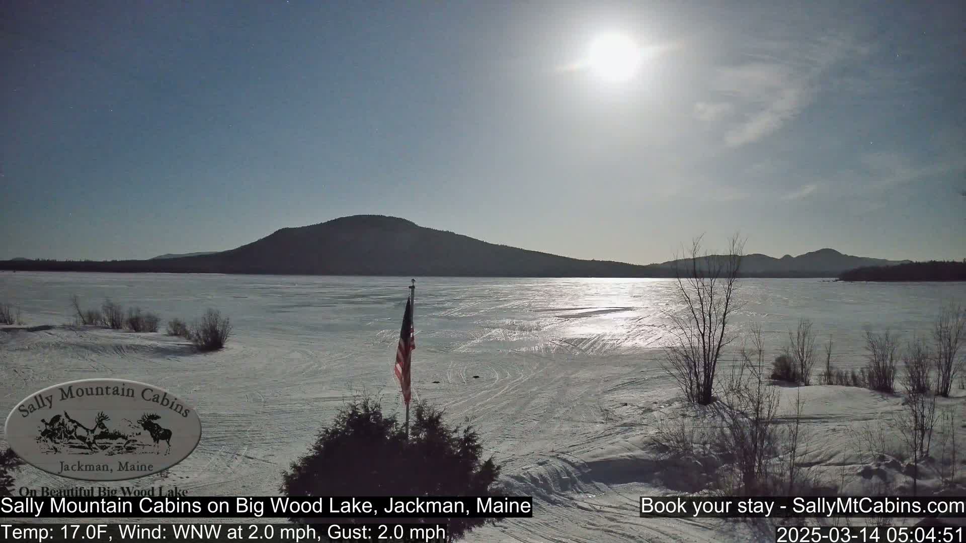 A frozen lake, under a partly cloudy sky, is visible with a distant mountain range and an American flag planted in the snow near the foreground.