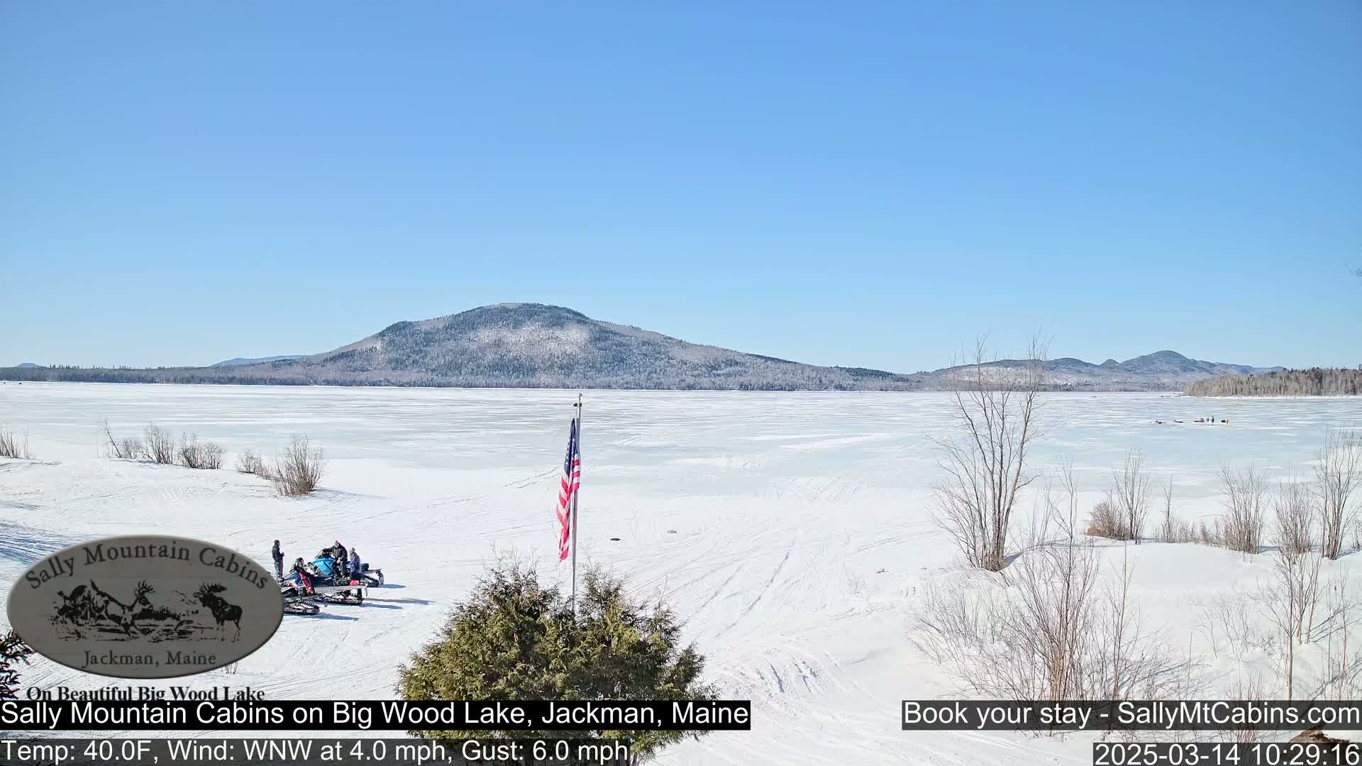 A snow-covered frozen lake stretches out to a distant mountain range under a clear blue sky, with several snowmobiles and people visible near the shoreline.