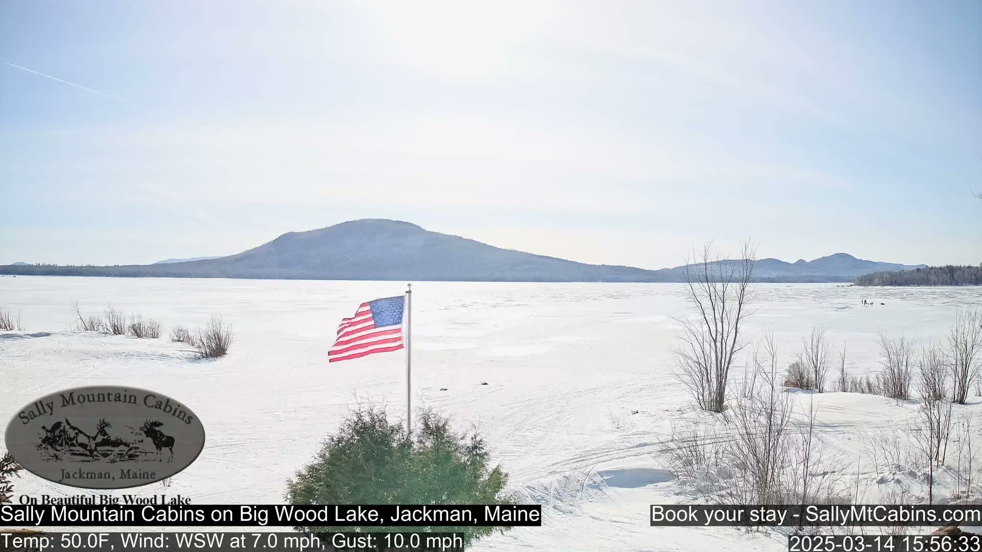 A frozen lake stretches out before a snow-covered shoreline, with a distant mountain range under a clear, bright sky, and an American flag waving in the breeze.