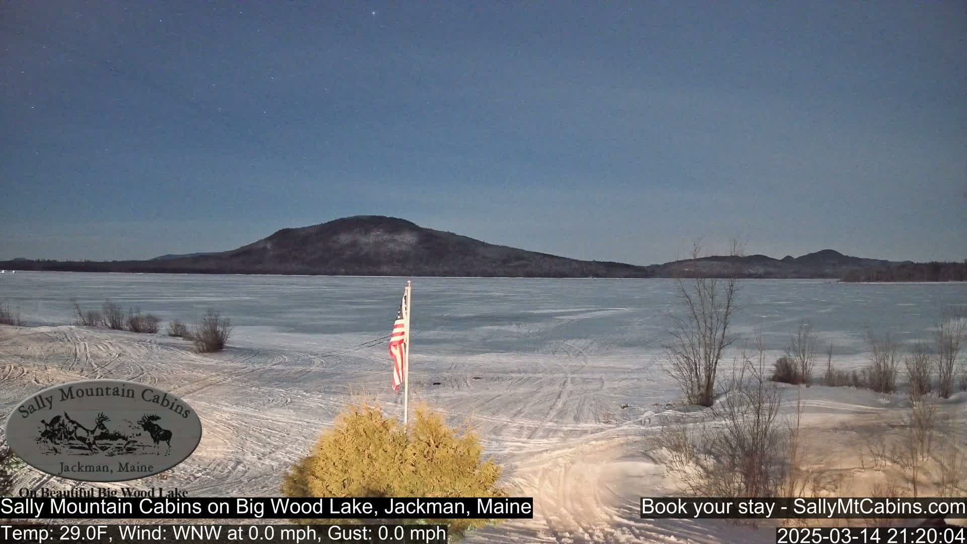 A frozen lake stretches before a snow-covered shoreline under a clear, dark night sky, with a distant mountain range visible and an American flag standing near the foreground.