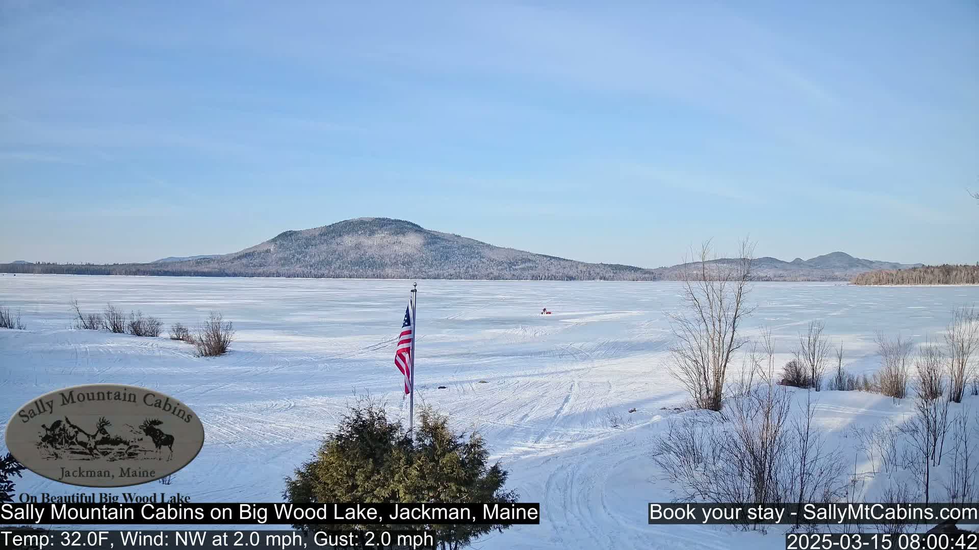 A frozen lake stretches to a snow-covered mountain under a clear blue sky, with a small American flag planted on the snowy shore.