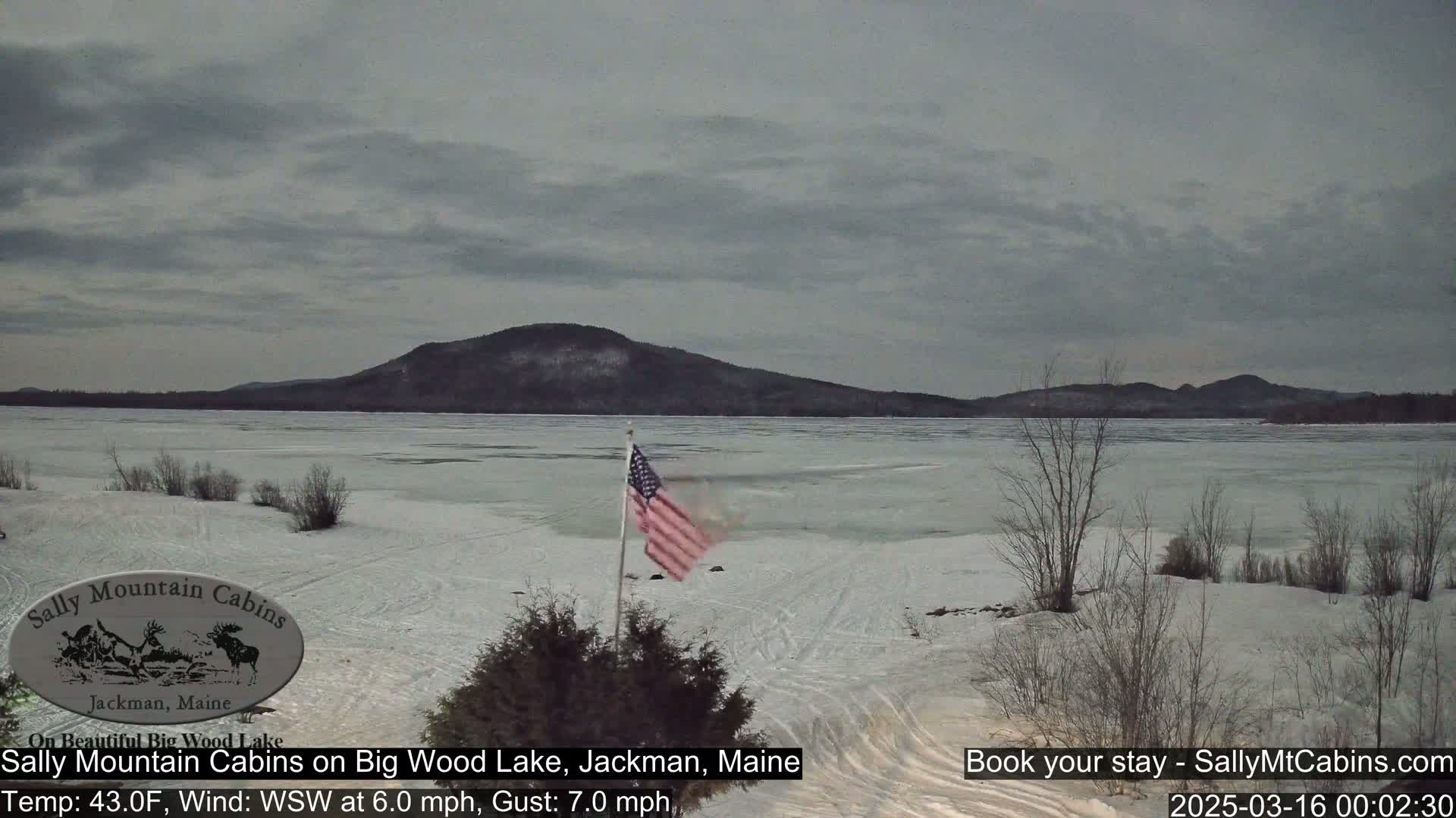 A partially frozen lake stretches to a snow-dusted mountain range under a cloudy sky, with an American flag waving in the foreground.