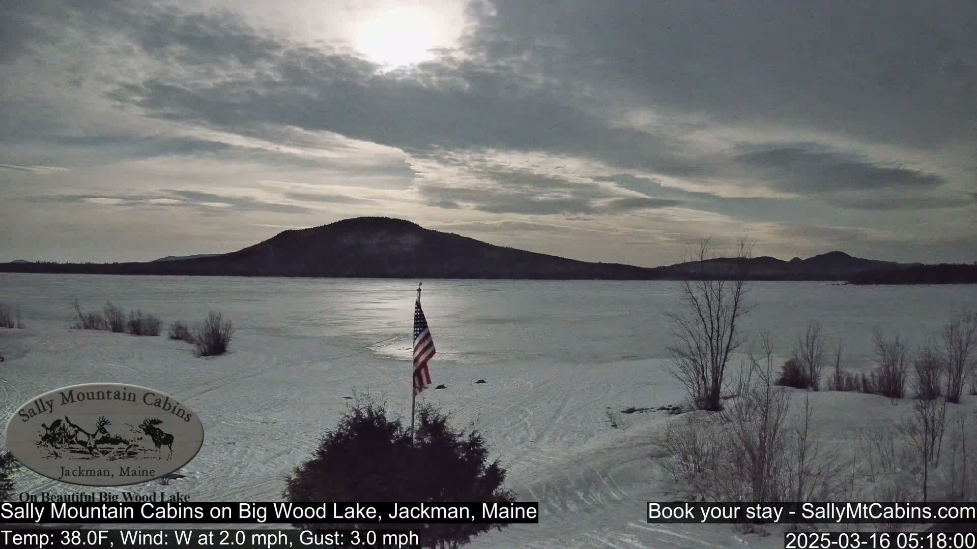 A frozen lake stretches to a distant mountain under a mostly cloudy sky, with a small American flag planted near the foreground.