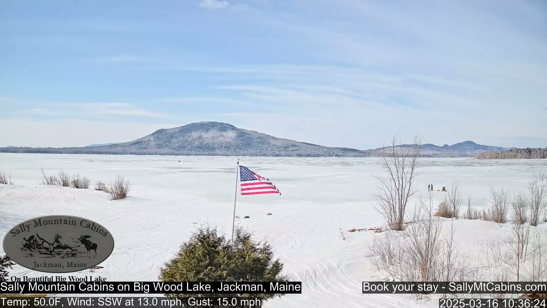 A frozen lake stretches to a snow-dusted mountain range under a mostly clear, bright sky; an American flag waves in the breeze from a lakeside flagpole.