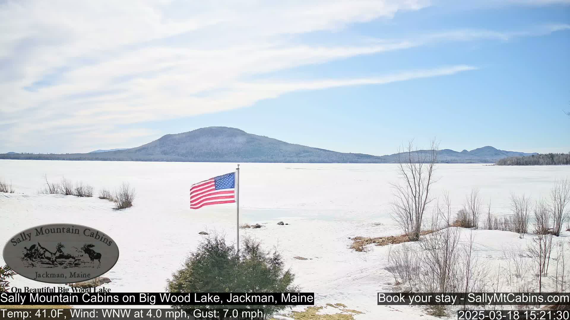 A snow-covered frozen lake stretches to a mountainous background under a mostly clear sky, with an American flag waving in the foreground.
