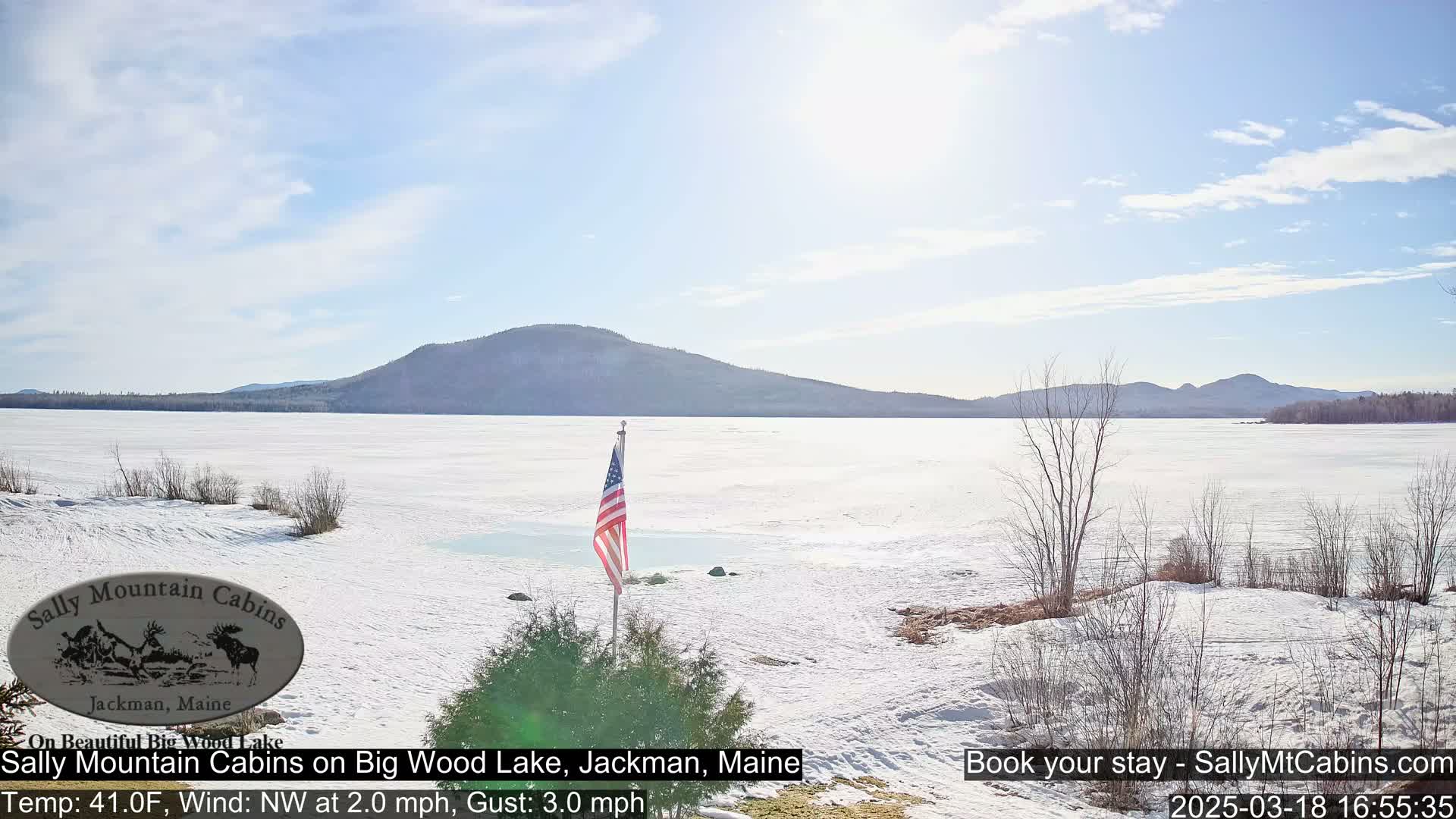 A partially frozen lake with snow-covered banks, a distant mountain range, and an American flag planted in the foreground under a mostly sunny sky.